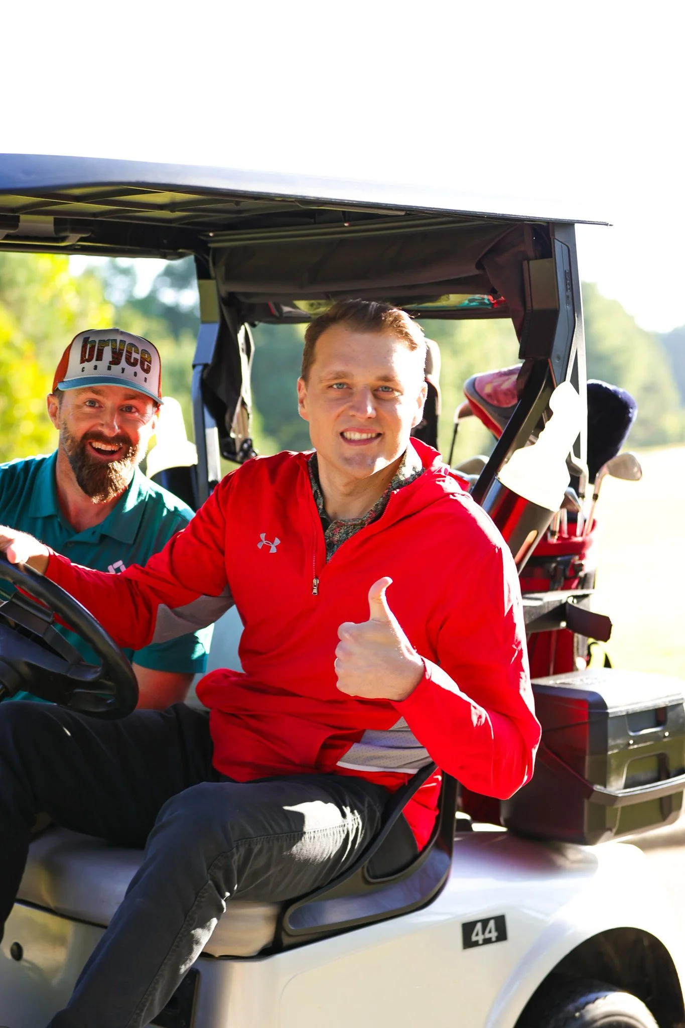 Two men sitting in a golf cart outdoors on a sunny day, one wearing a red jacket giving a thumbs up and the other wearing a teal shirt and a cap, smiling.
