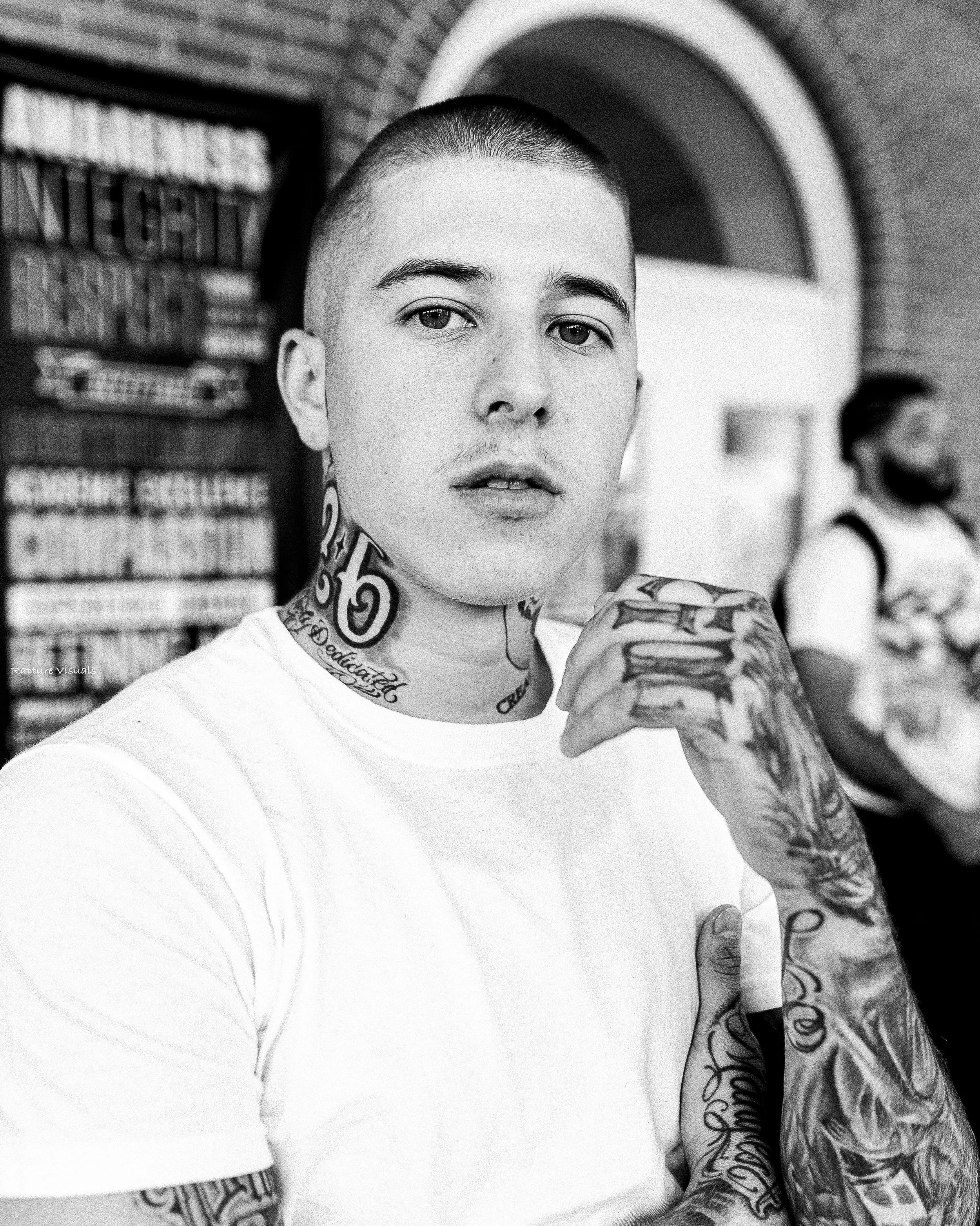 Black and white photo of a tattooed young man with a buzz cut hairstyle, with hand up near his face, taking a selfie in an urban setting. Background includes brick walls and a man in a face mask, out of focus.