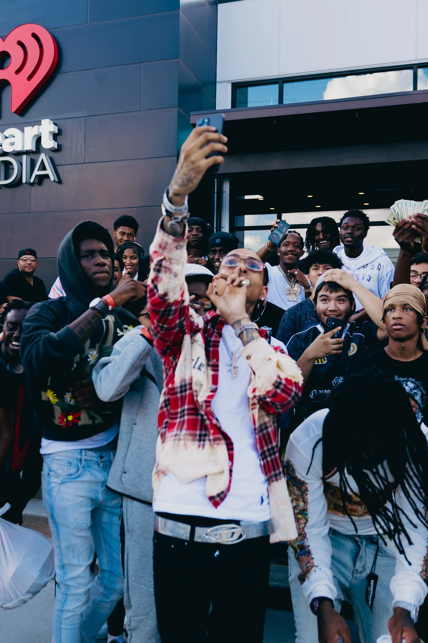 A group of young people taking a selfie outside a building with a sign that says 'art MEDIA'. The group includes several males and females, some holding money, and others looking at the camera and smiling.