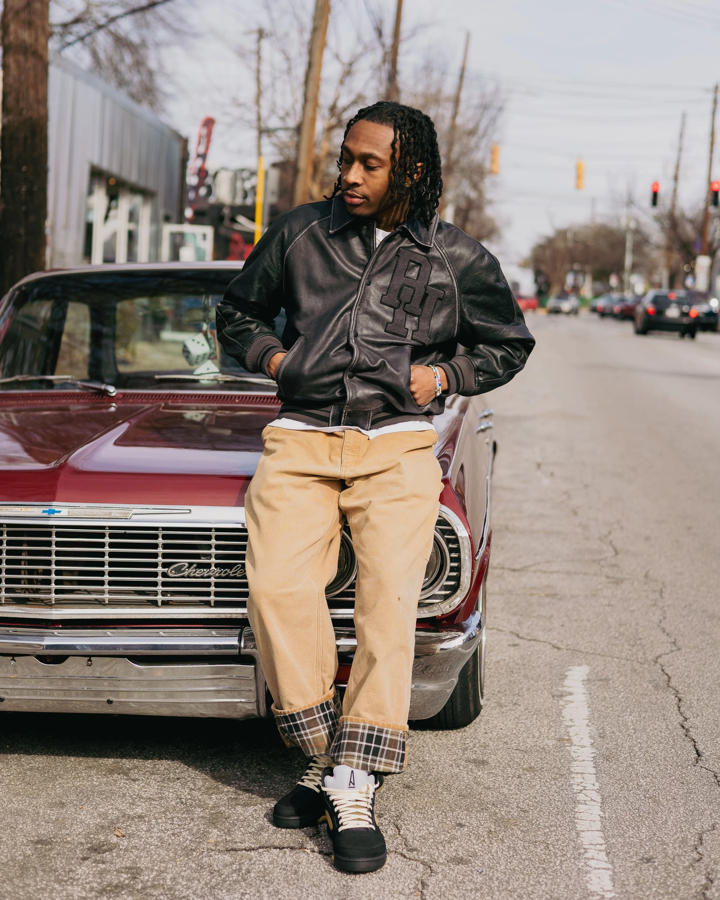 A young man with dreadlocks leaning against the hood of a vintage maroon Chevrolet car in a parking lot. He is wearing a black leather varsity jacket, tan pants with plaid cuffs, and black and white sneakers. Background includes leafless trees, utili