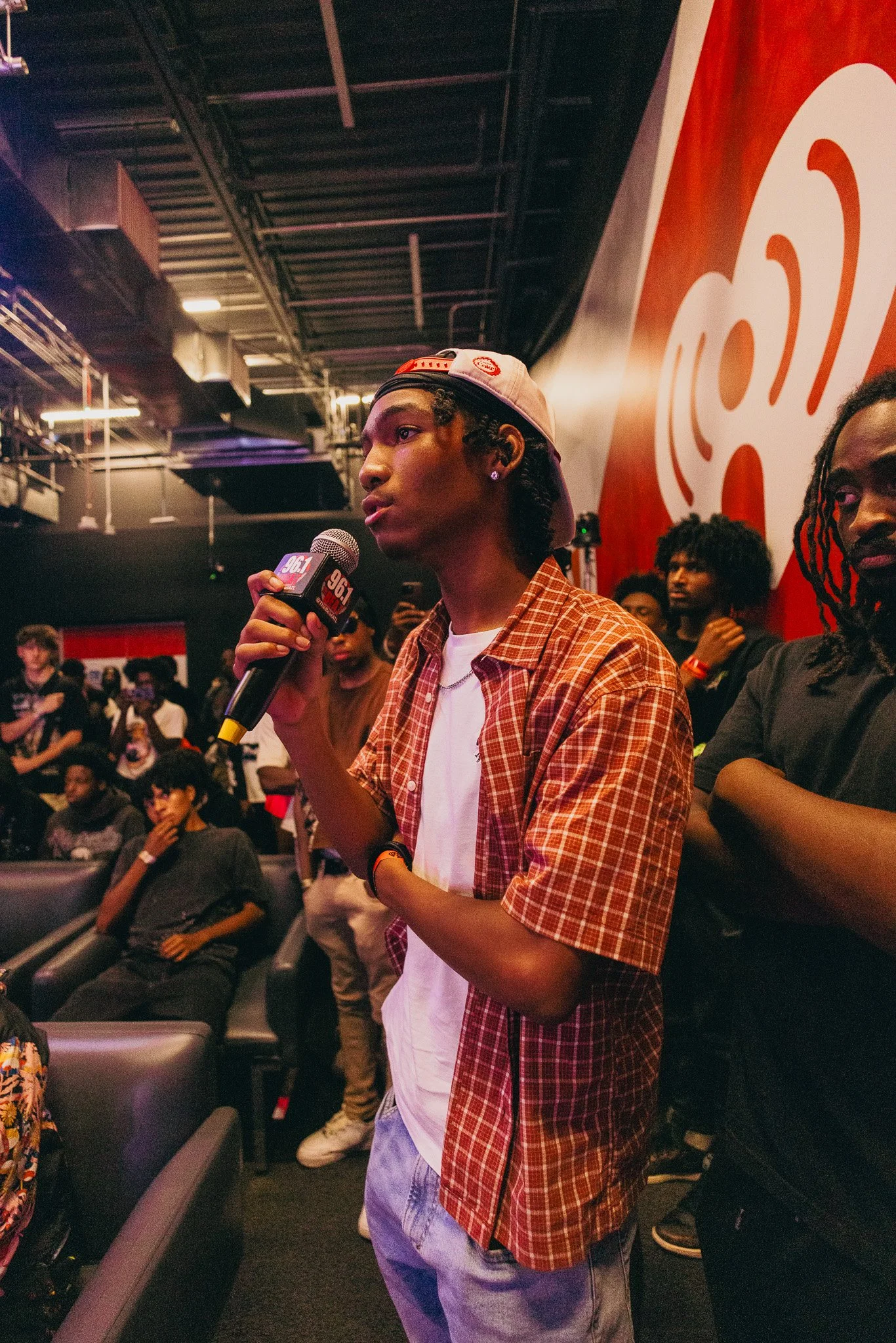 A young man with a white baseball cap worn backwards, earrings, and a checkered shirt, holding a microphone with a radio station logo during an event at a crowded indoor venue with a red and white sign in the background.