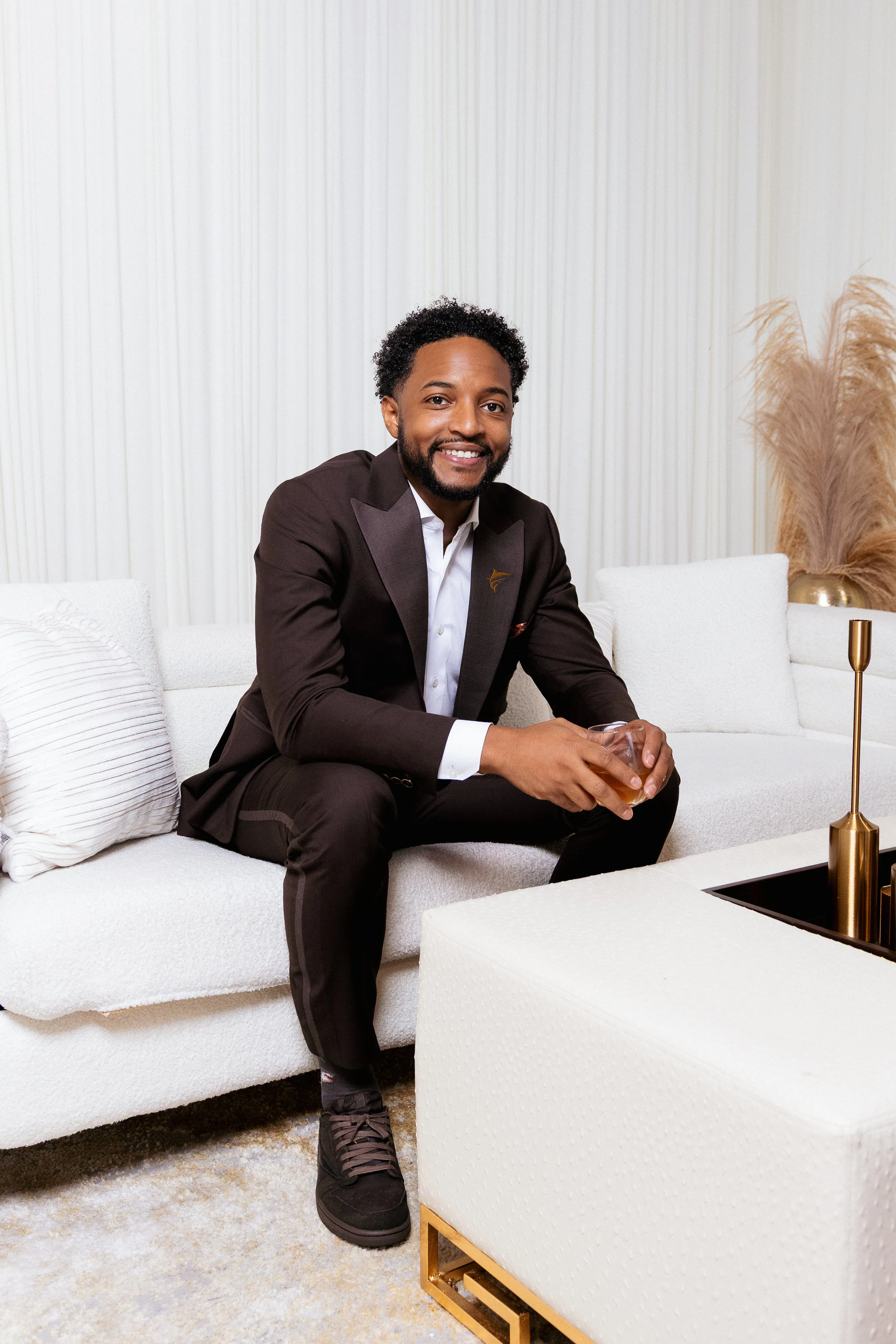 A man with curly black hair and a beard, wearing a dark brown suit and white shirt, sitting on a white sofa with a drink in his hand, smiling at the camera in a modern, minimalist living room.