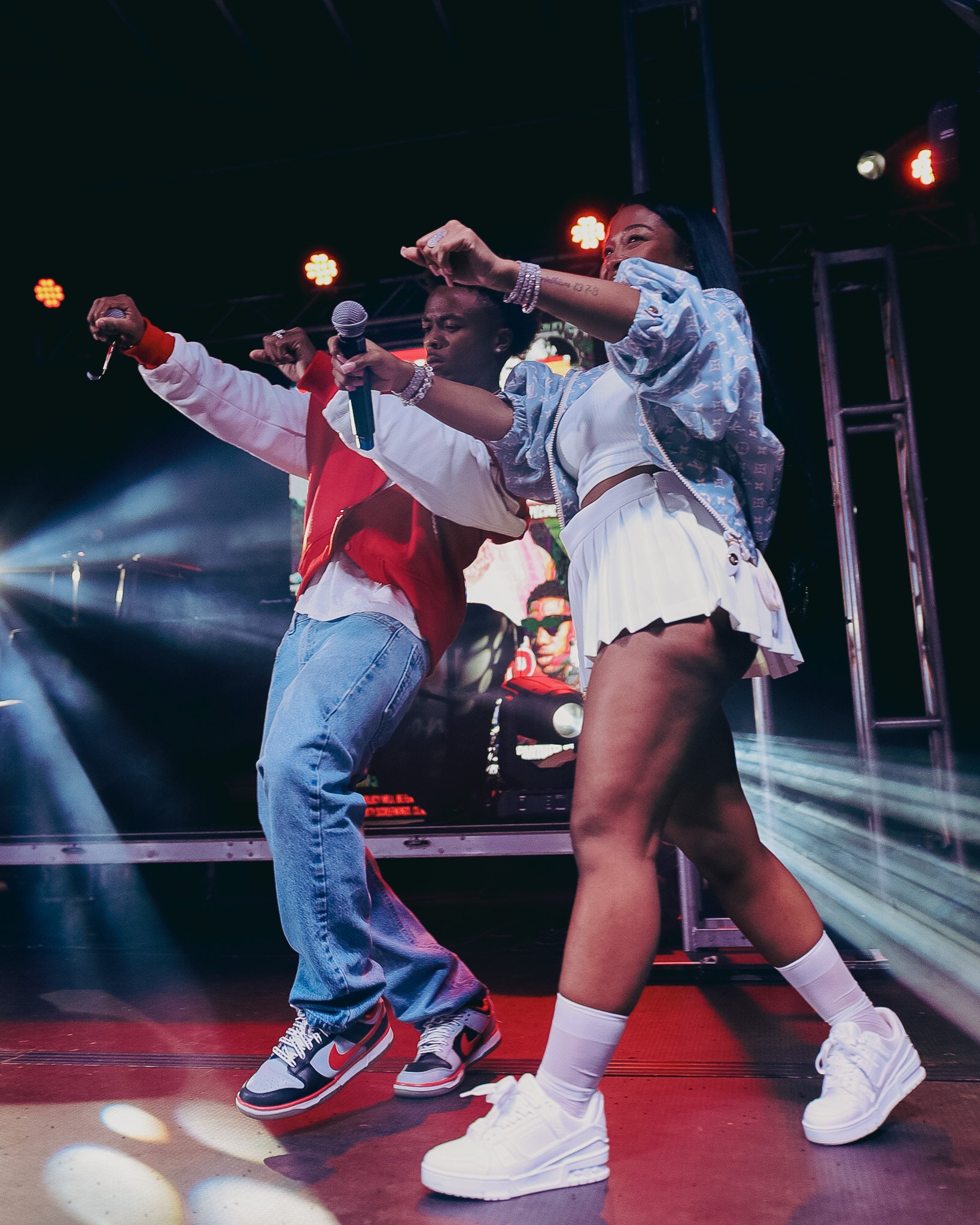 Two women dancing on stage with colorful lights shining down, one holding a microphone, wearing casual clothing and sneakers, the other in sportswear with a mask.