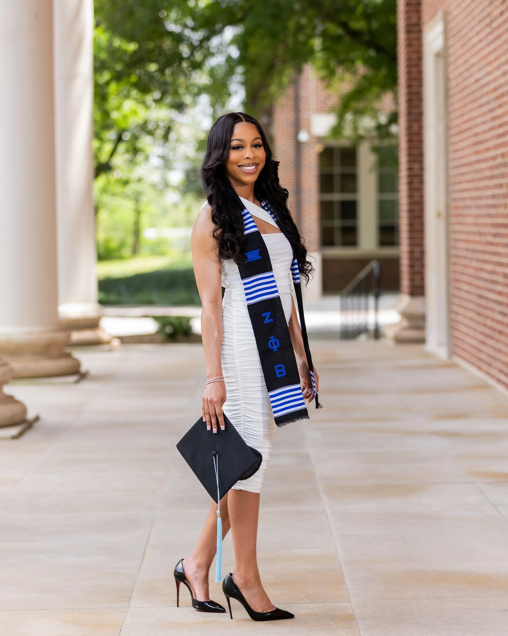 A woman dressed in a white dress and black high heels, wearing a graduation stole, holding a black diploma cover, standing outdoors on a sidewalk near a brick building with green trees in the background.