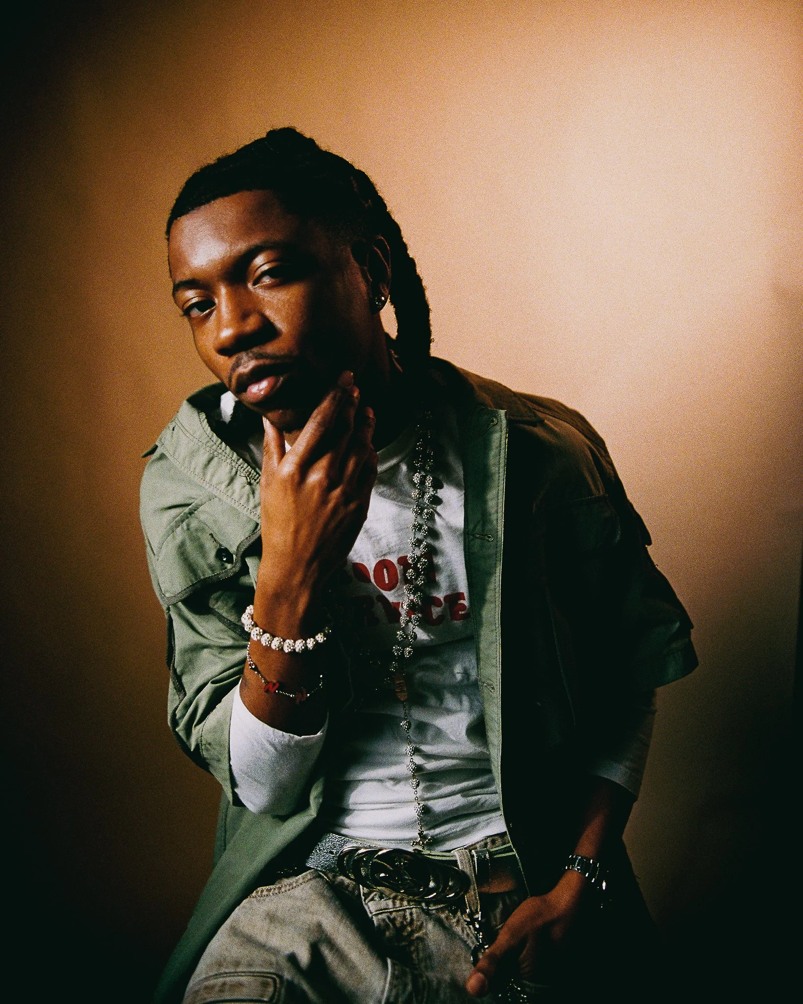 Portrait of a young man with dark skin and dreadlocks, wearing a green jacket, white shirt, and jewelry, posing with hand on chin against a plain beige background.