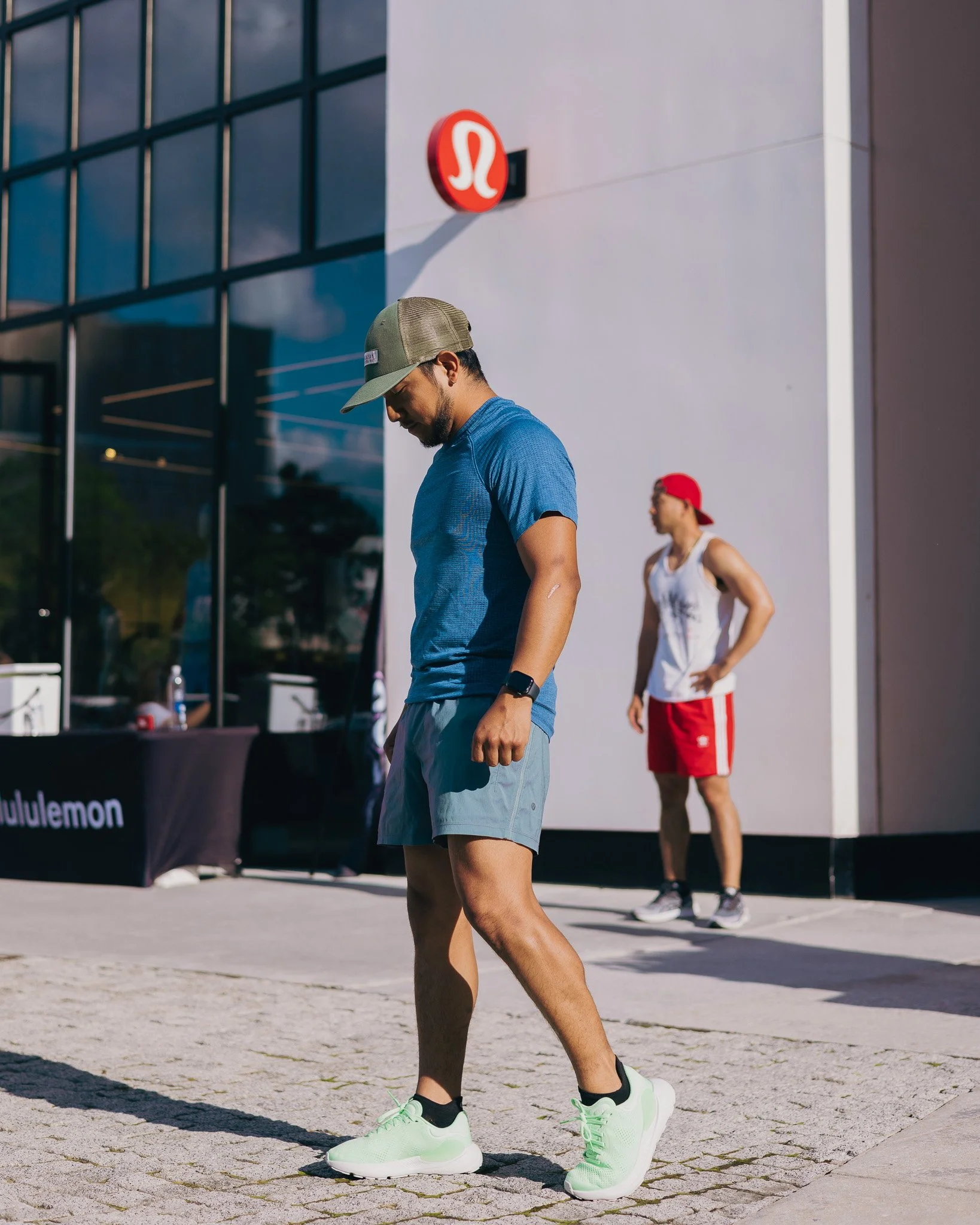 A man in a blue athletic shirt and light gray shorts bending over in a parking lot, wearing a beige cap, black smartwatch, and lime green running shoes. In the background, a woman in a white tank top and red shorts, with a red cap, is walking.