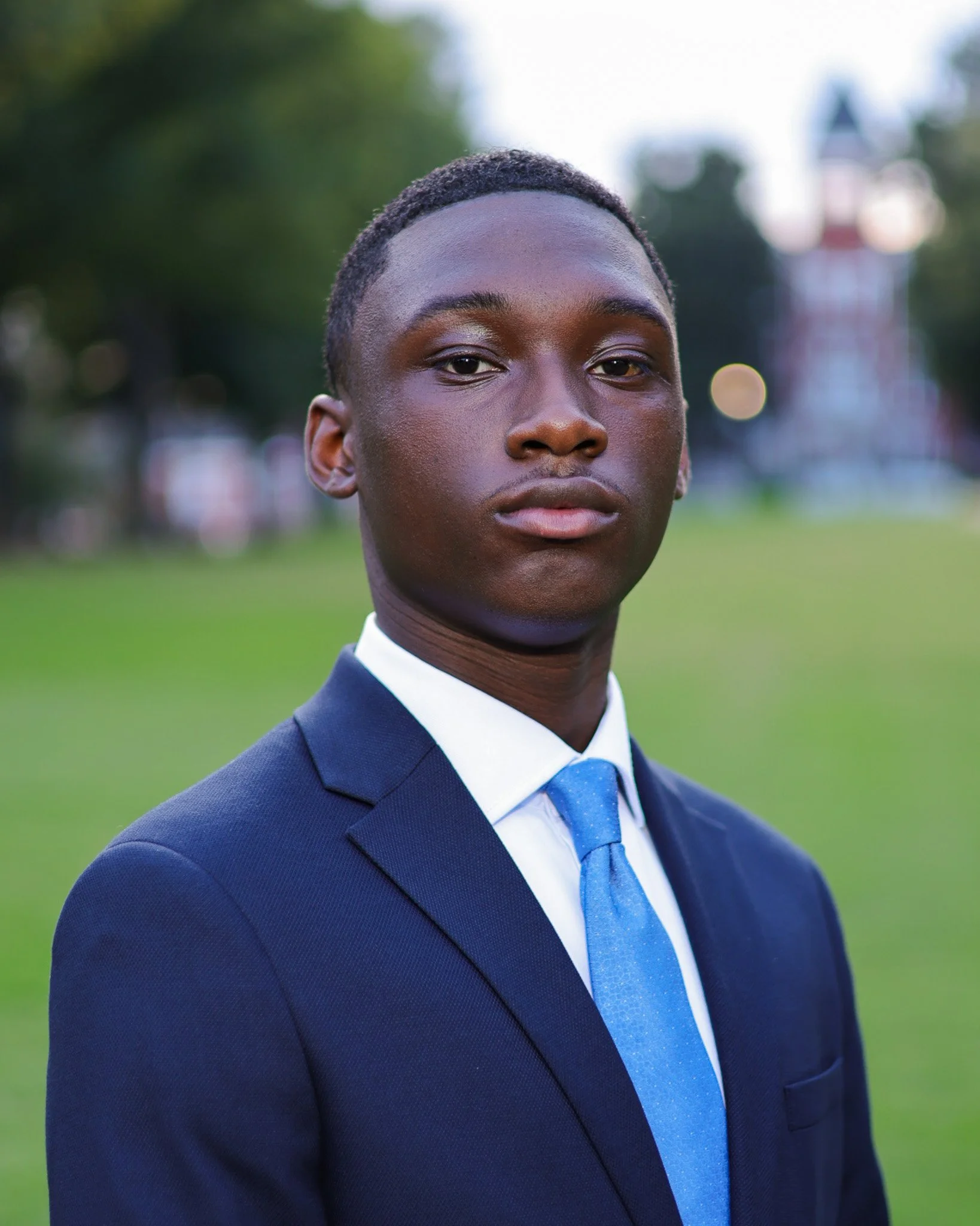 A young African American man in a navy blue suit, white shirt, and light blue tie, standing outdoors with a blurred background of green trees and a distant lighthouse.