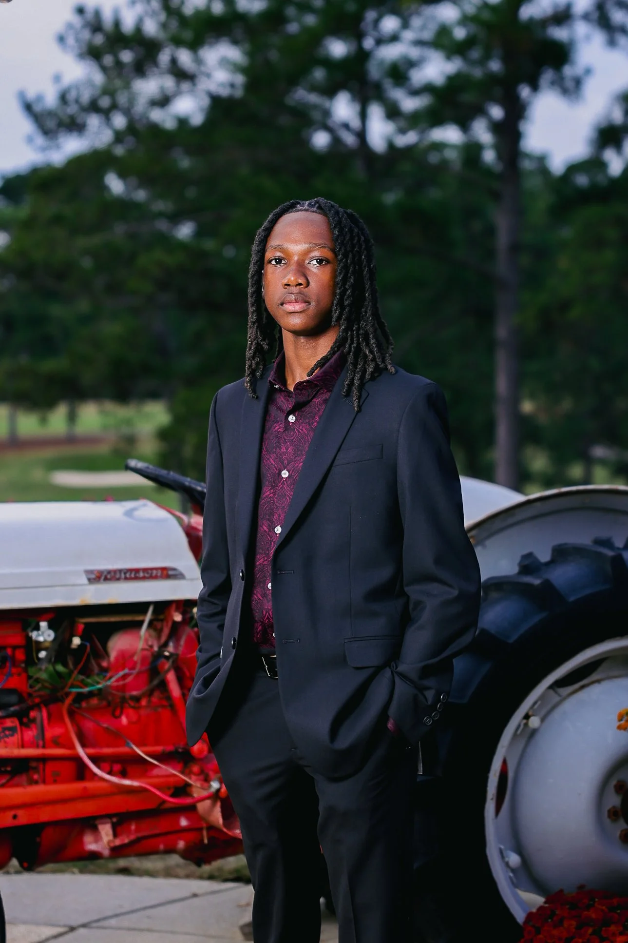 A young man in a dark suit and patterned shirt standing outdoors with vintage tractors and greenery in the background.