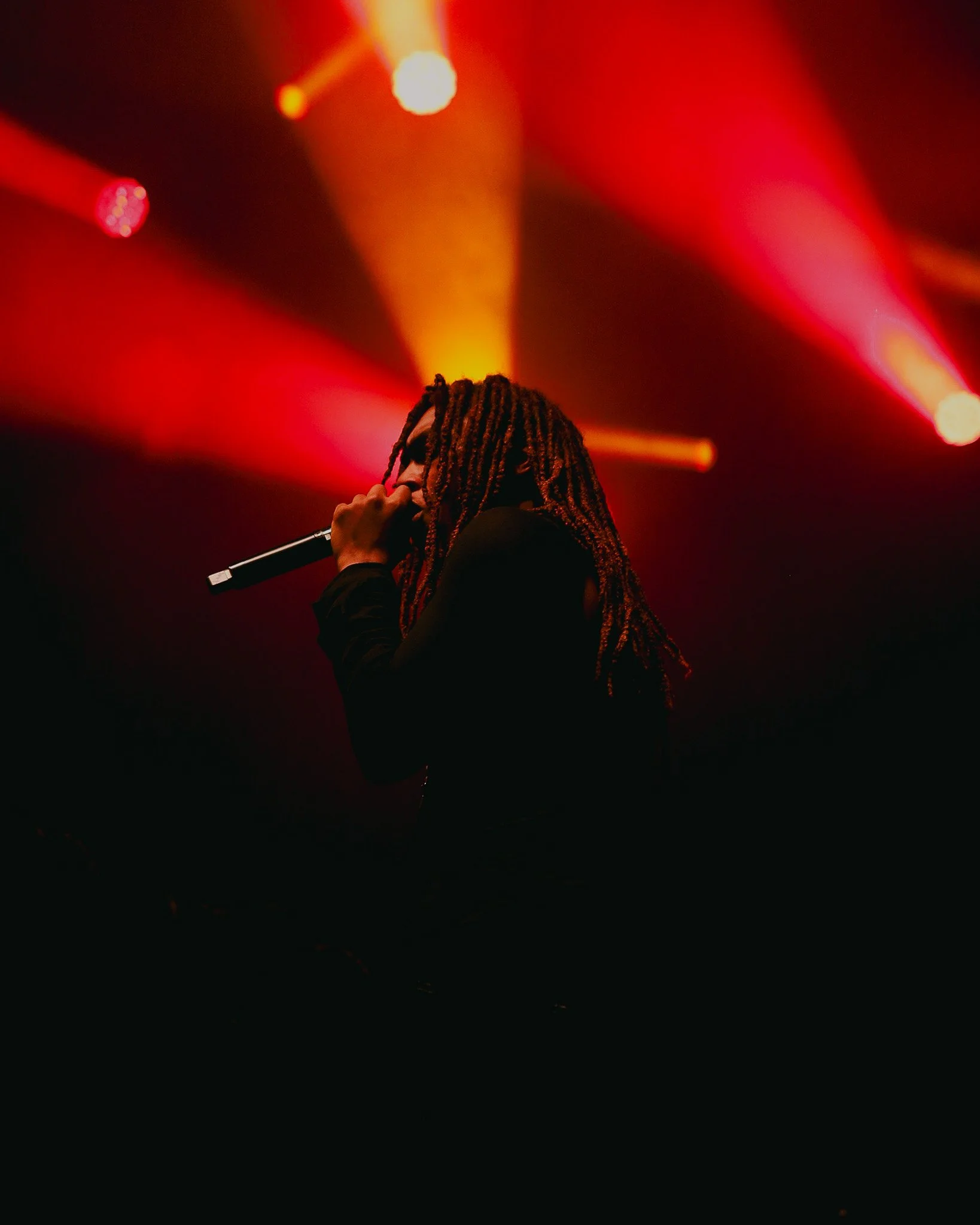 Person with braided hair holding a microphone, performing on stage with colorful stage lights in the background.