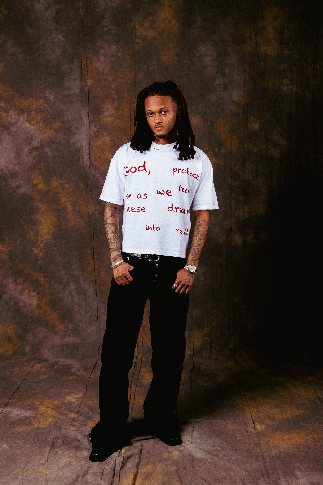 A young man with dreadlocks and tattoos wearing a white t-shirt with red writing, black pants, and a watch, standing against a dark, textured background.
