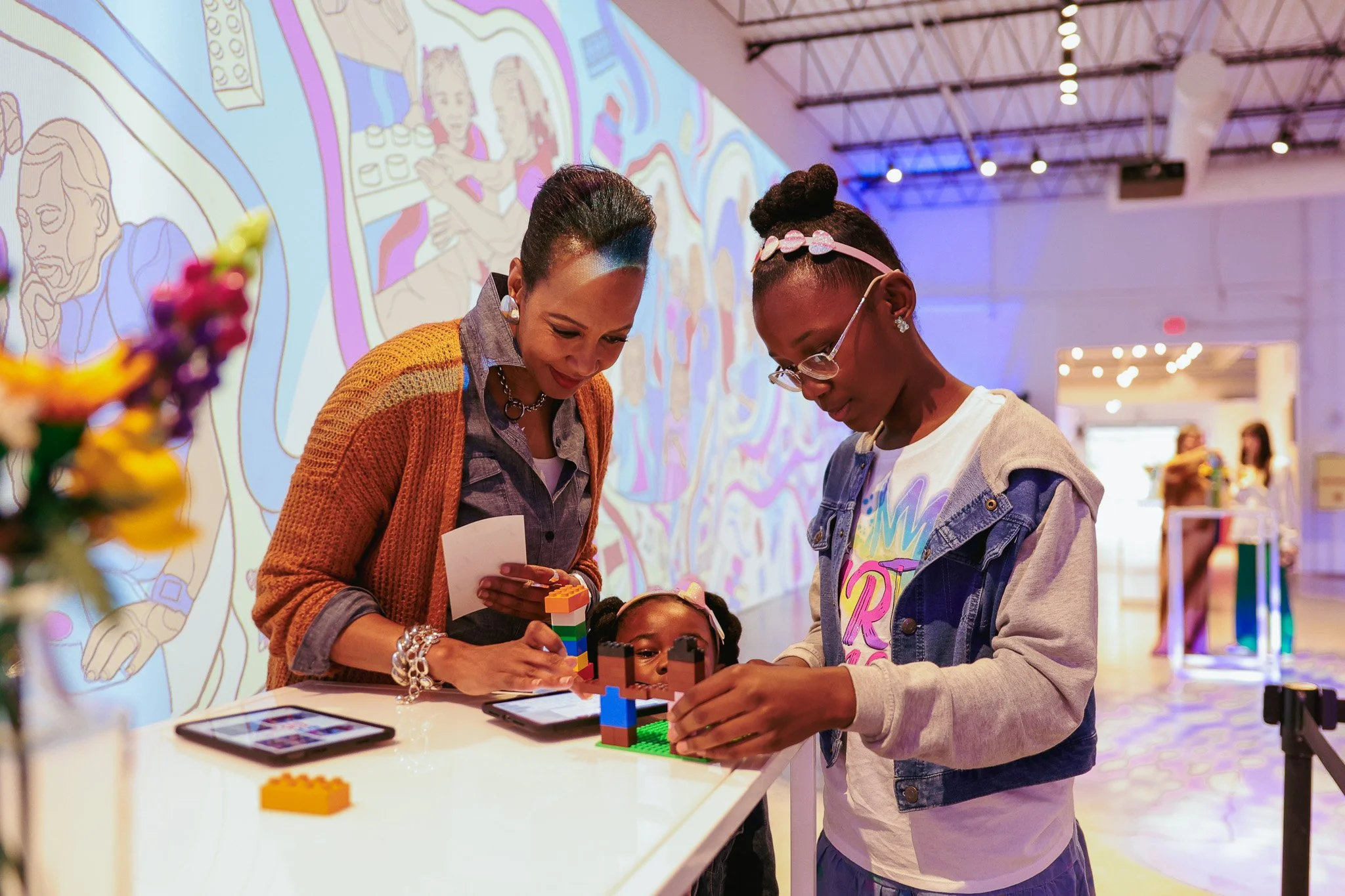 Two women and a girl engaging in a LEGO building activity at an indoor event, with colorful wall art in the background.