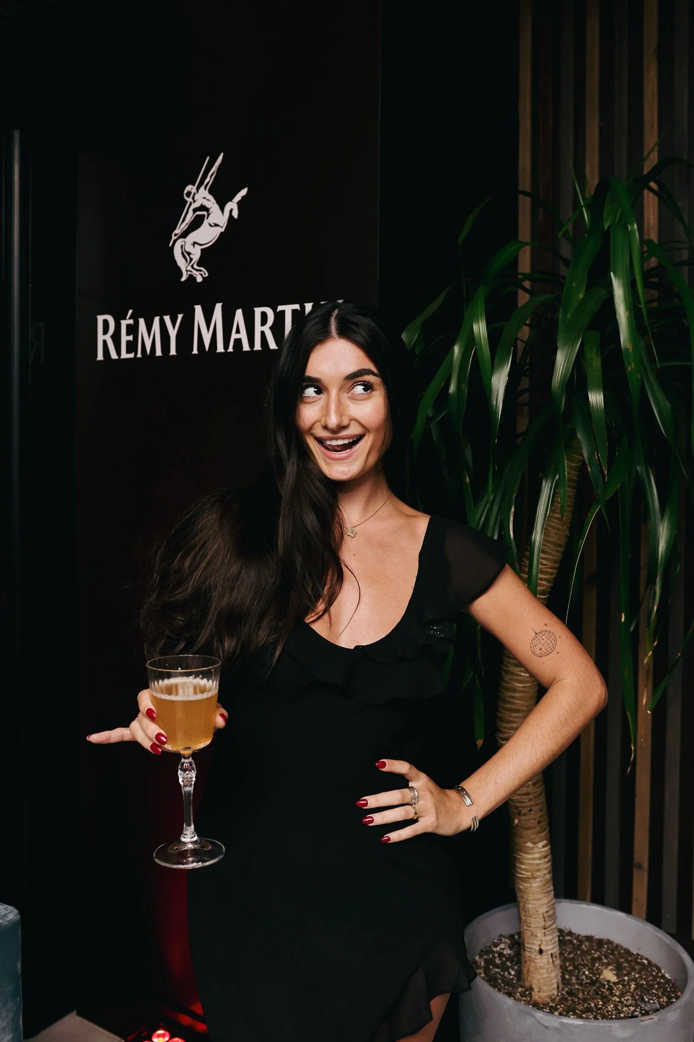 A woman with long dark hair holding a glass of beer, smiling and looking to her right, standing next to a large potted plant with a tall stem and leaves. She has a tattoo on her left forearm and is wearing rings and red nail polish. Behind her is a b