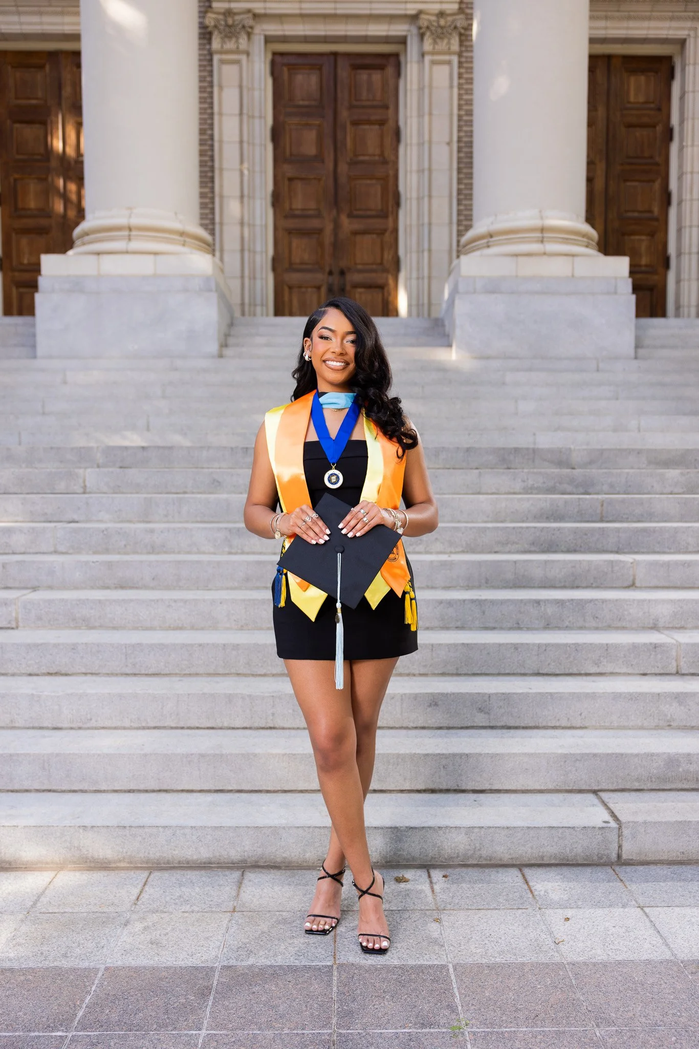 A woman in a black dress and high heels is standing on the stairs outside a building, wearing graduation regalia including a stole, medallion, and cap.