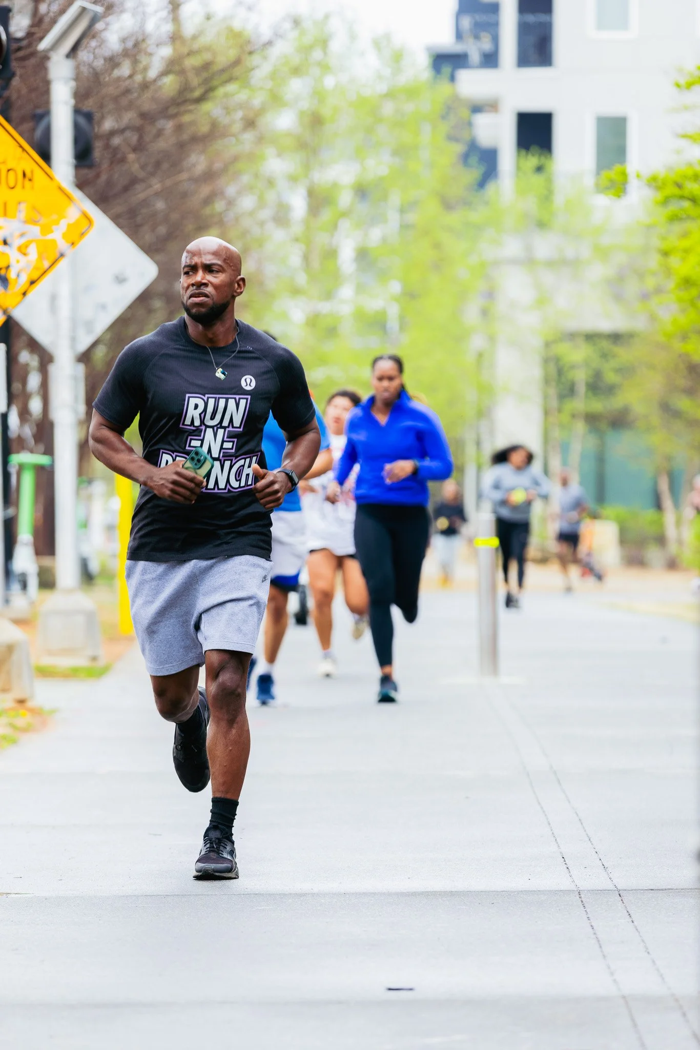 A man running outdoors on a city sidewalk, wearing a black t-shirt with the words 'Run the Punch' and gray shorts.