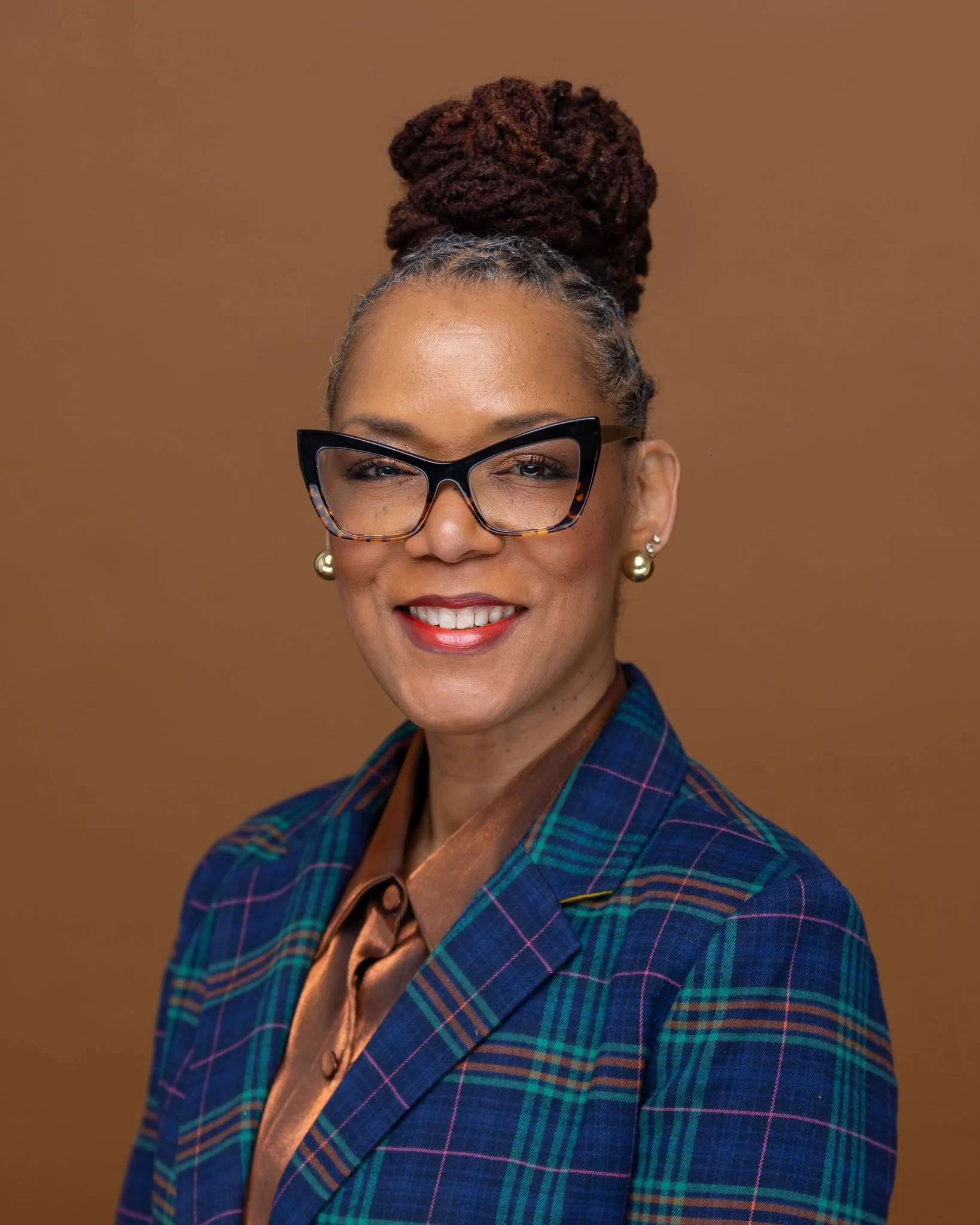 A woman with glasses, pearl earrings, and a hairstyle with a high bun of dreadlocks, wearing a plaid blazer and a brown blouse, smiling against a plain brown background.