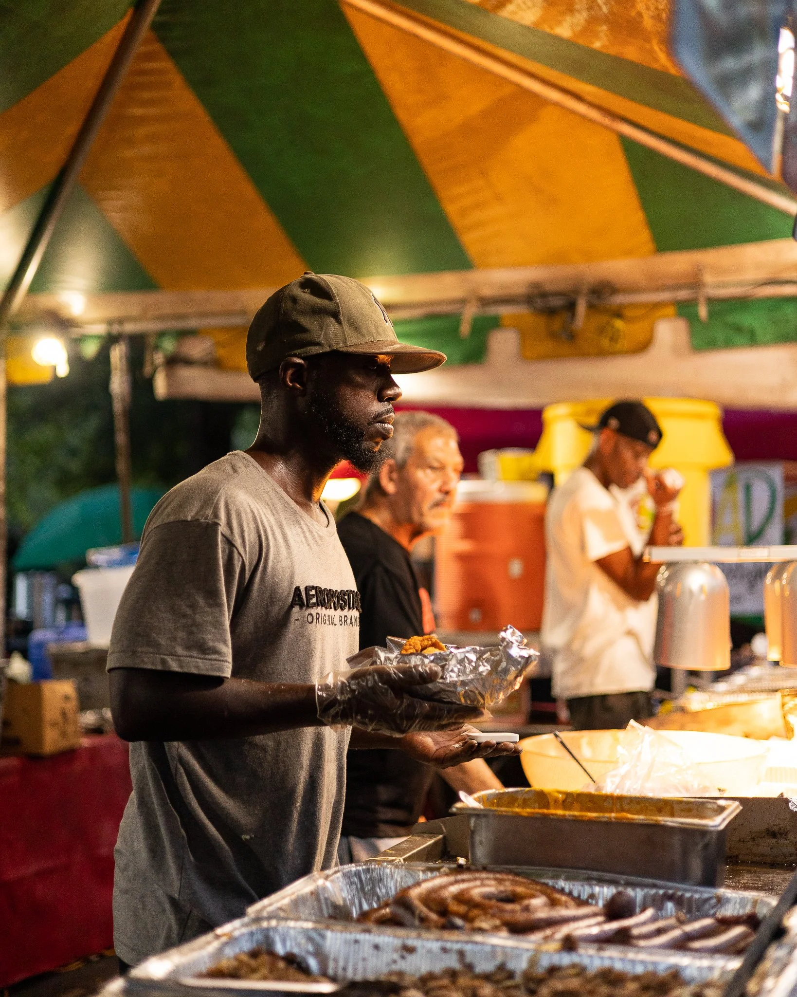 A man wearing a gray t-shirt and a camouflage baseball cap holding a tray of fried food at an outdoor food stand at night, with others preparing food in the background under a colorful tent canopy.