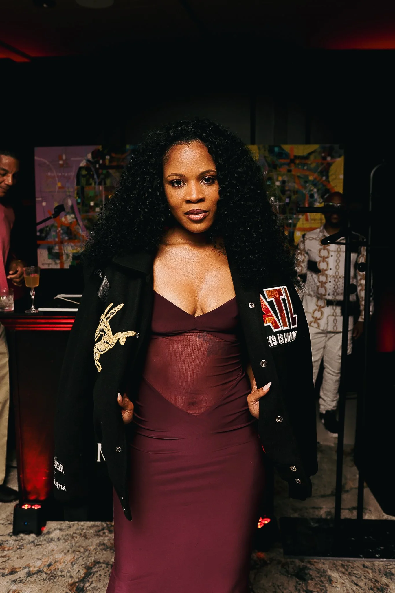 A woman with long, curly black hair wearing a burgundy dress and a black jacket with patches, standing in front of a dark background with colorful abstract artwork. She has her hands on her hips and is looking at the camera.