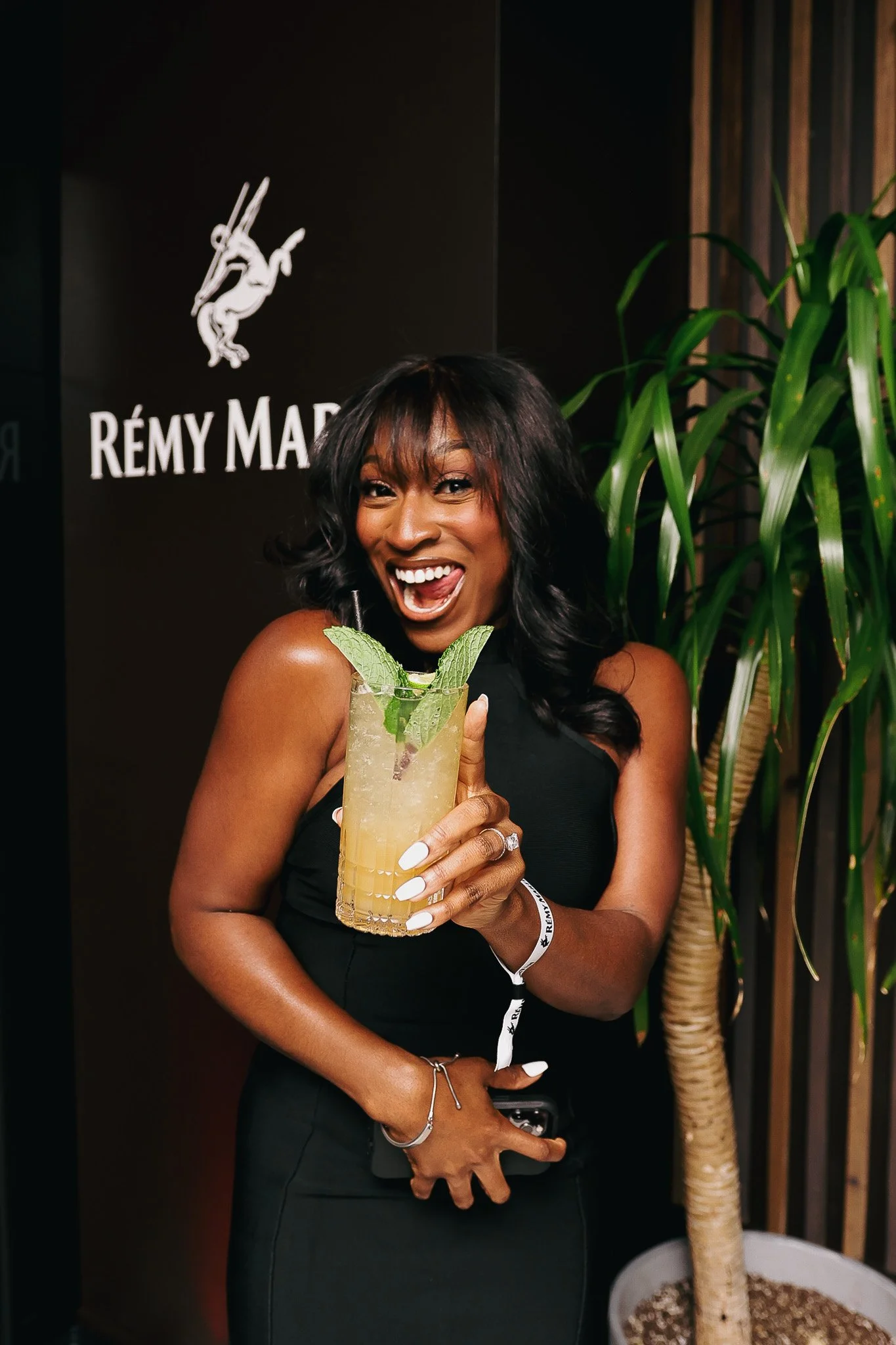 A smiling woman holding a lemon mojito cocktail garnished with mint, standing in front of a black wall with a Remy Martin logo and a green potted plant.