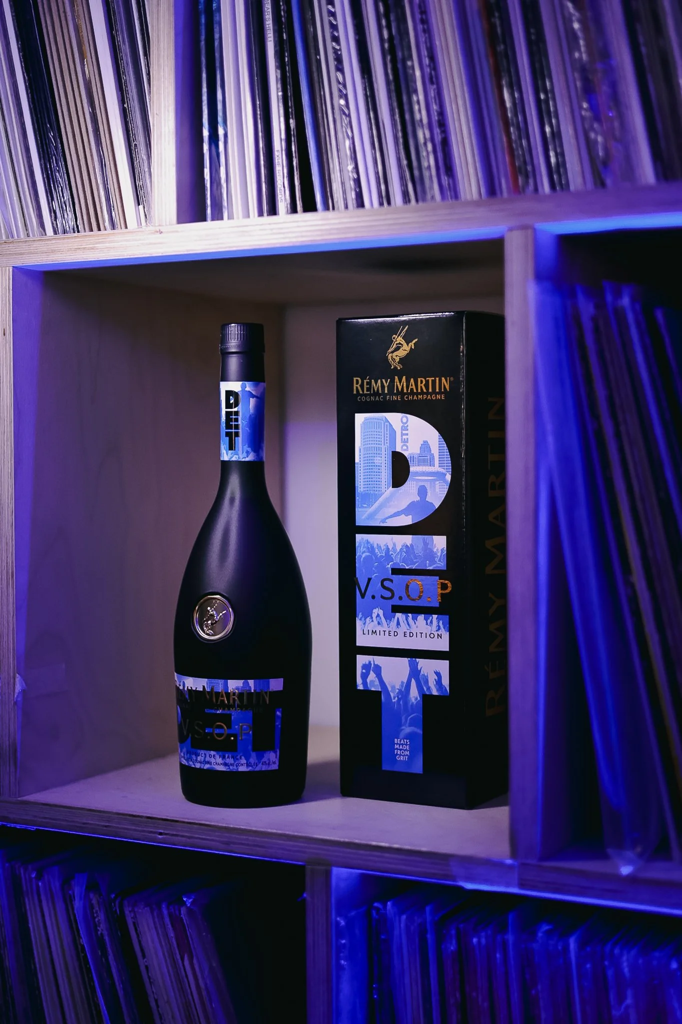 A black bottle of Rémy Martin V.S.O.P. limited edition cognac with a black box next to it, both placed inside a wooden shelf with a collection of vinyl records on the upper and lower shelves, illuminated by purple and blue lighting.