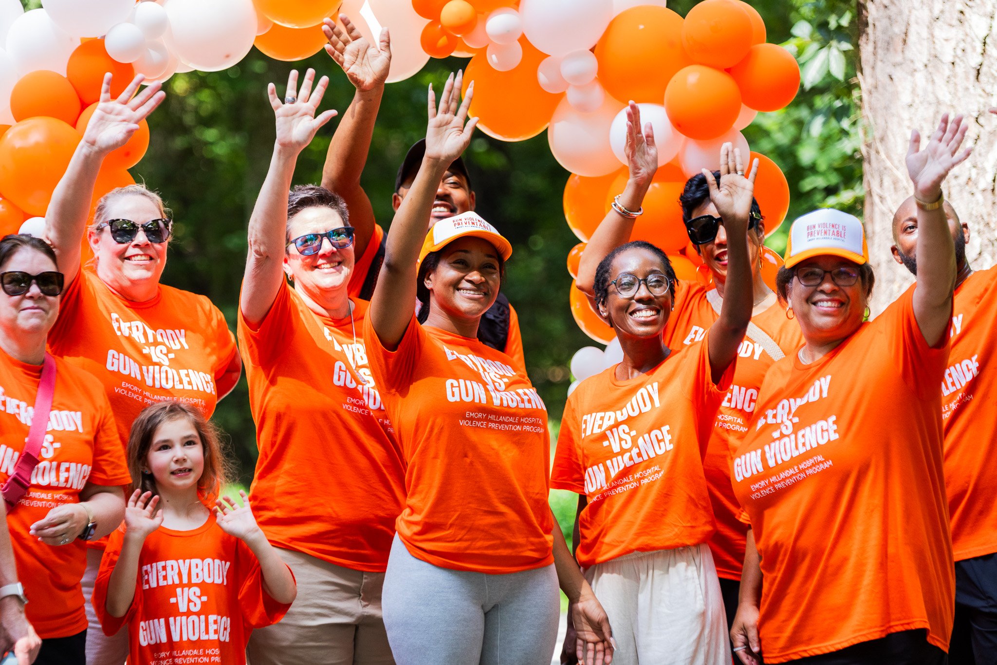 Group of diverse people wearing orange shirts and hats, waving, at an outdoor event with orange and white balloons overhead.