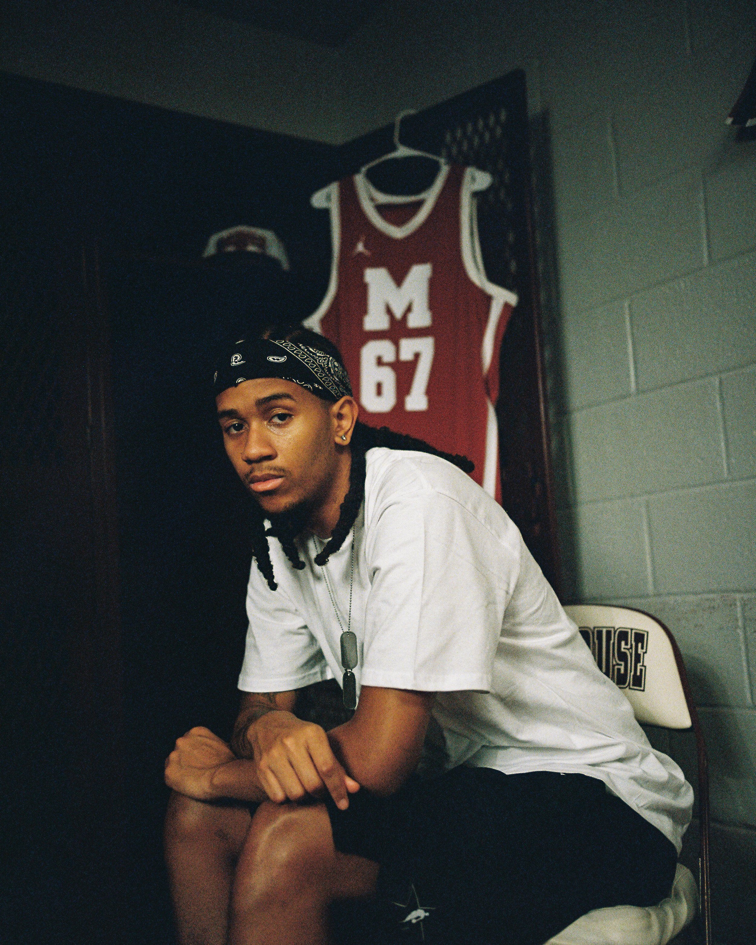 Young man with a bandana, wearing a white t-shirt, sitting in a dimly lit room with a basketball jersey hanging on the wall behind him.
