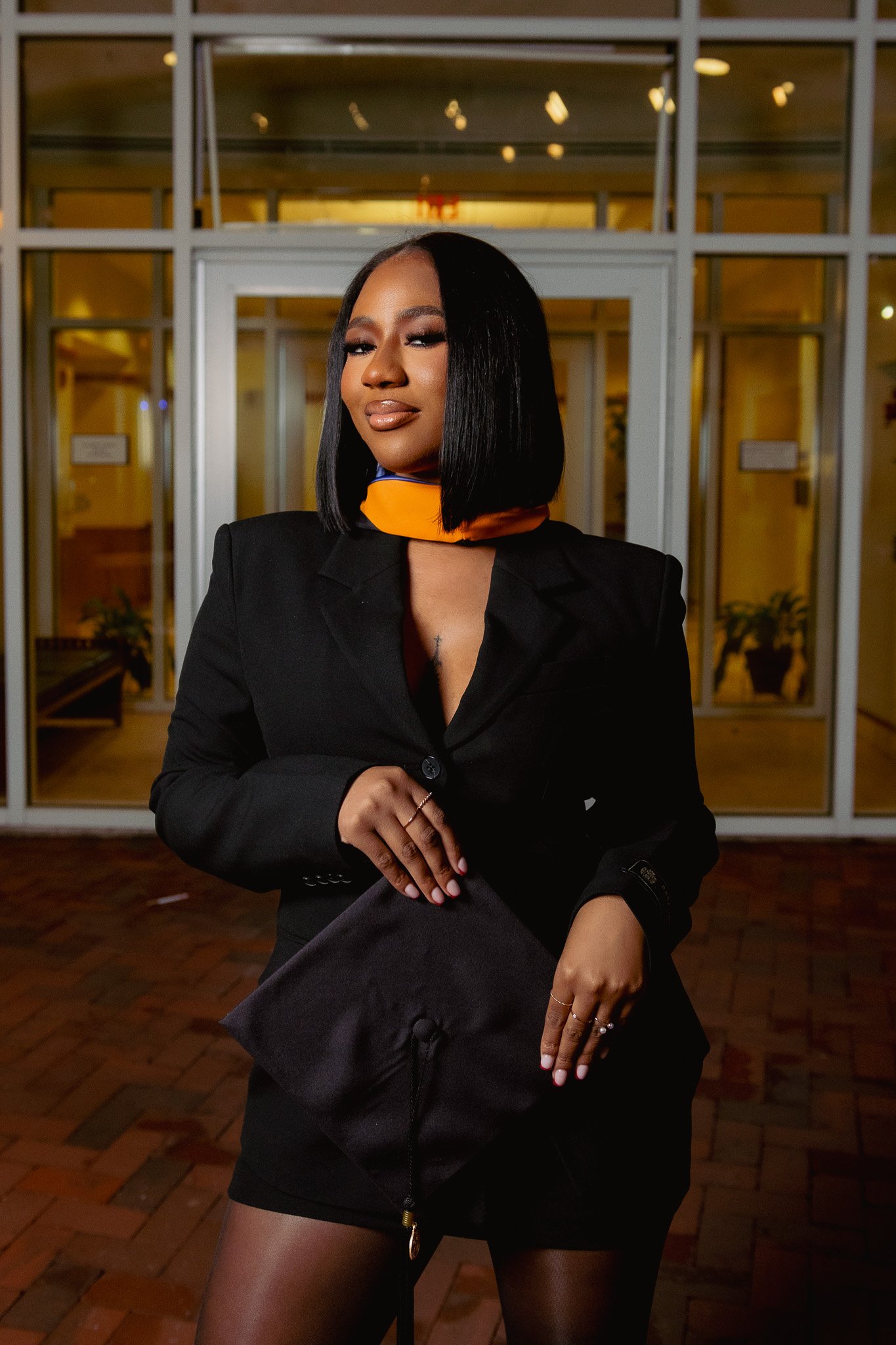 A woman in a black blazer and black skirt standing indoors in front of glass doors, holding a black bag, wearing sheer black tights, with a confident expression and a slight smirk.