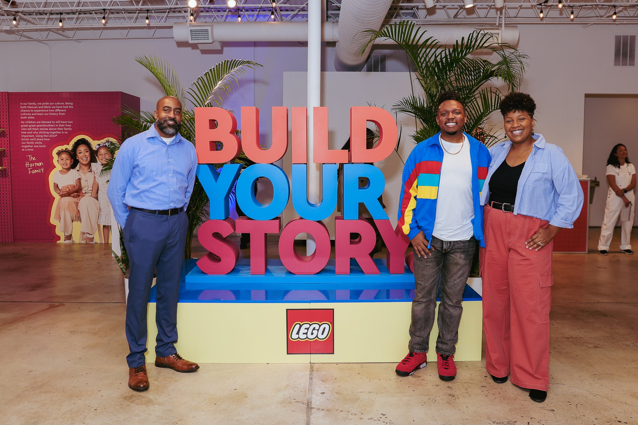 Three people standing in front of a large pink and blue sign that says 'BUILD YOUR STORY' with a LEGO logo below it. They are indoors, with green plants and other people in the background.