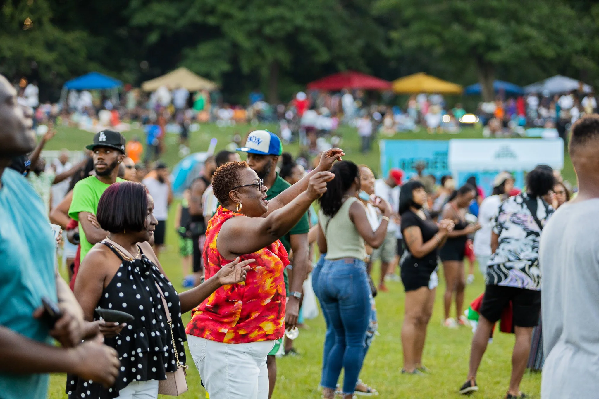 A diverse group of people attending an outdoor event in a park, with tents in the background and trees.
