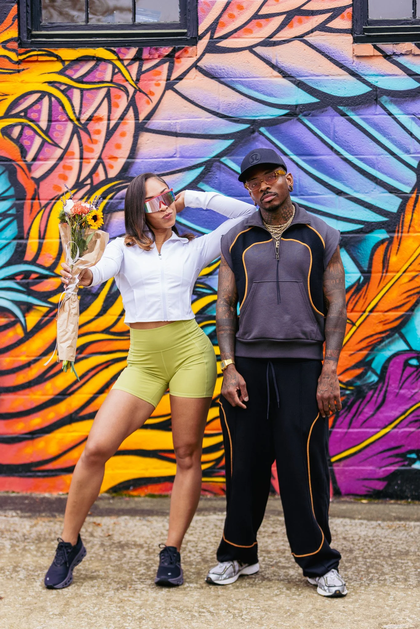 A woman and a man standing in front of a colorful graffiti mural. The woman is holding a bouquet of flowers, wearing sunglasses, a white zip-up jacket, yellow shorts, and sneakers. The man is wearing a black cap, orange glasses, a sleeveless black ho