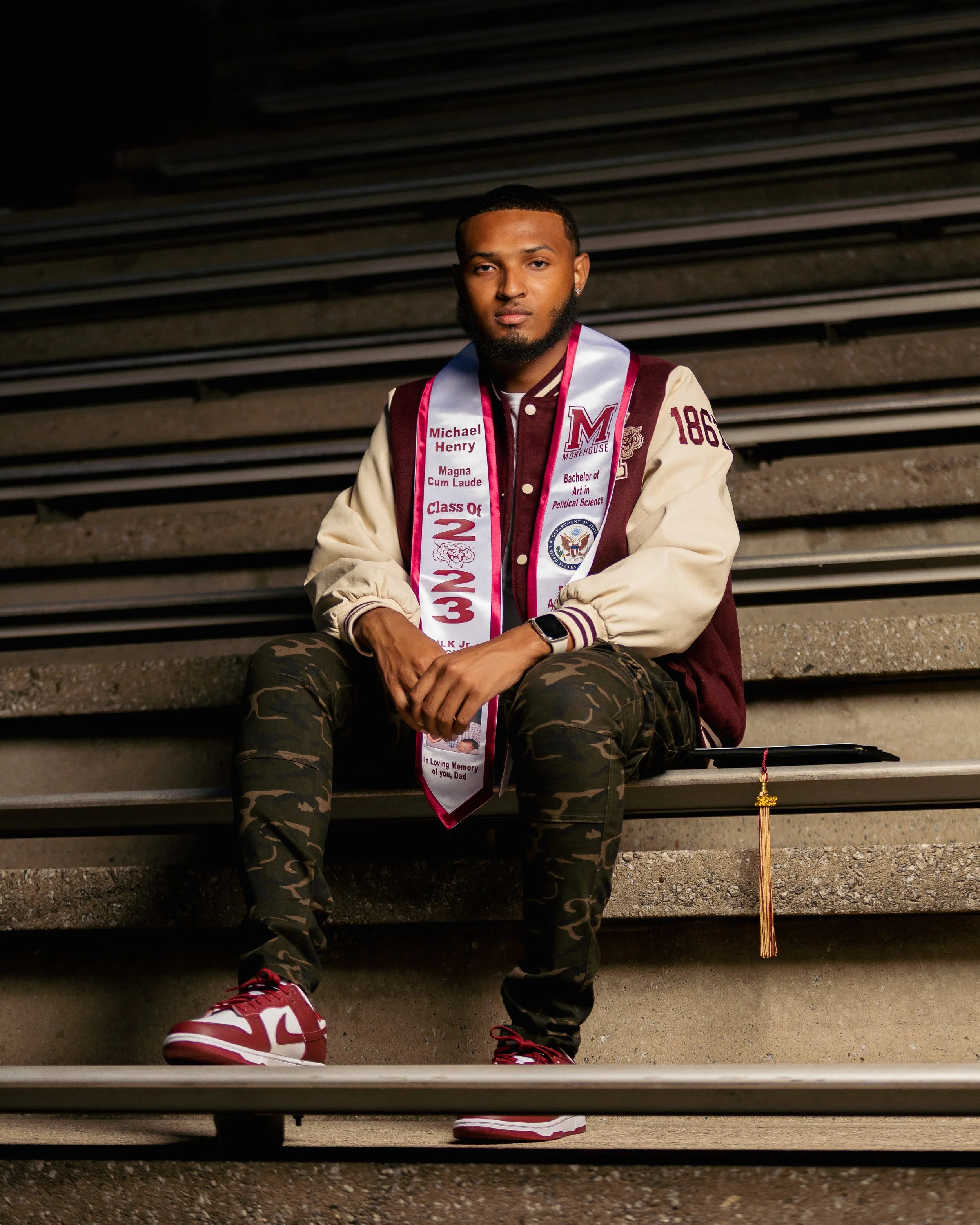 Young man sitting on stadium stairs wearing a graduation gown and sash, sporting a beard, camo pants, and red and white sneakers.