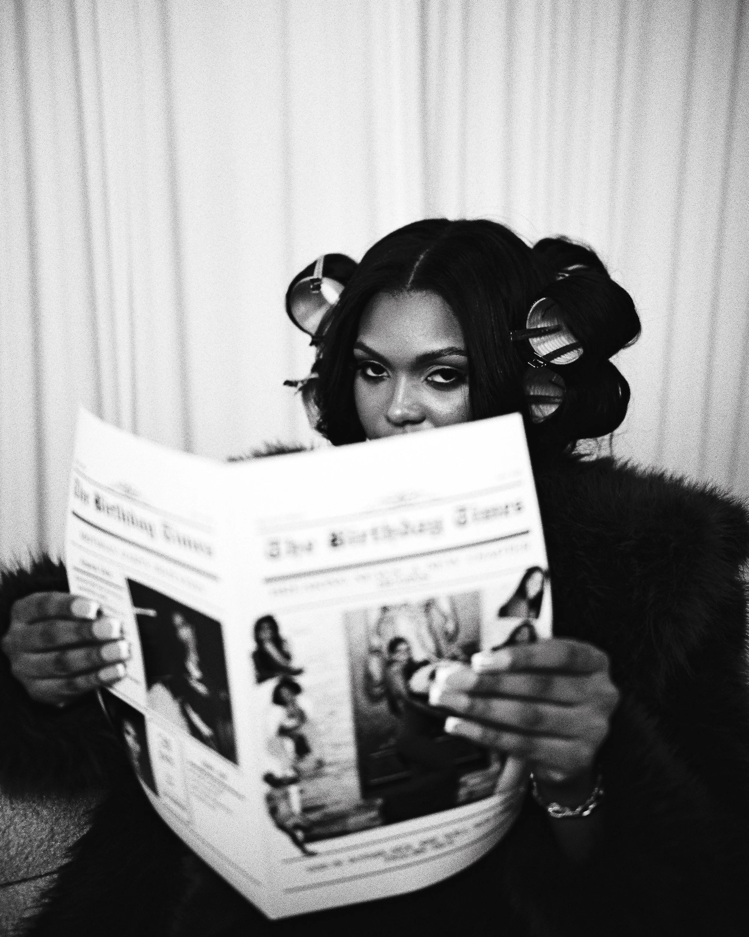 A woman with dark hair in rollers looking into a mirror and reading a magazine.