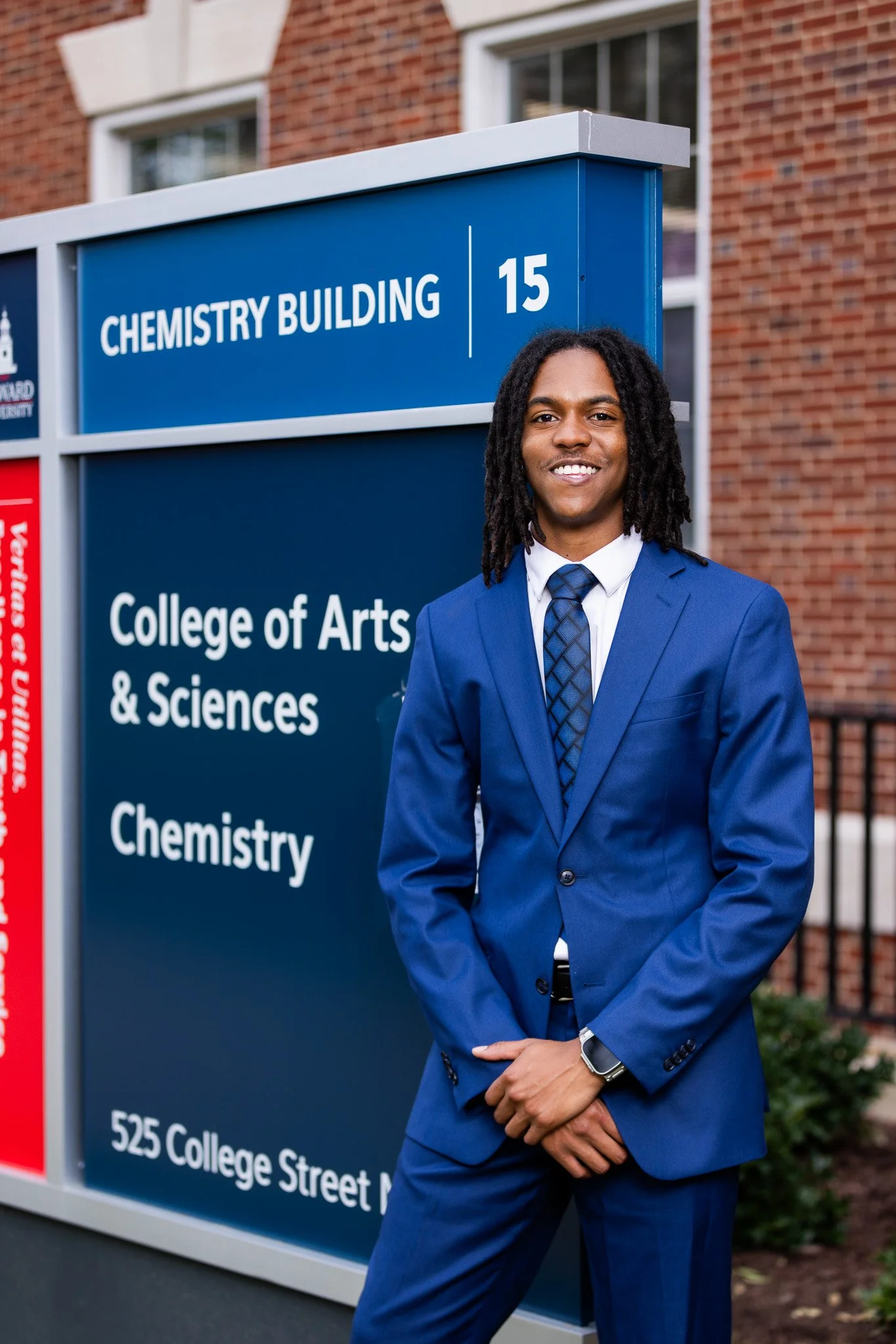 A young man in a blue suit and patterned tie smiling and standing with arms crossed in front of a university sign that reads 'Chemistry Building 15, College of Arts & Sciences, 525 College Street.' The background features a brick wall and windows.