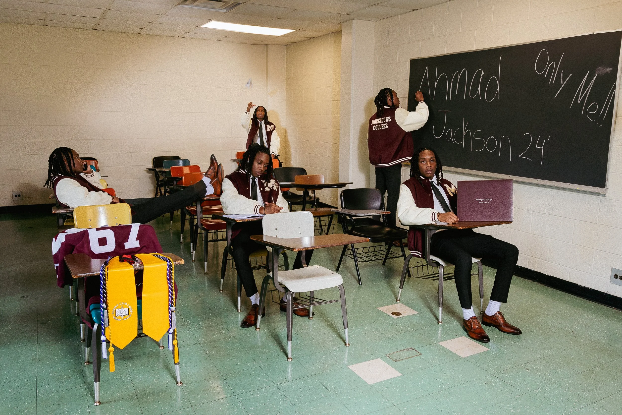 A classroom with students wearing burgundy and white jackets, some sitting at desks and one standing at a chalkboard writing. The blackboard has the words 'Amadi', 'Jackson 24', and 'Only Mom.' The classroom has a green tile floor, a drop ceiling, an