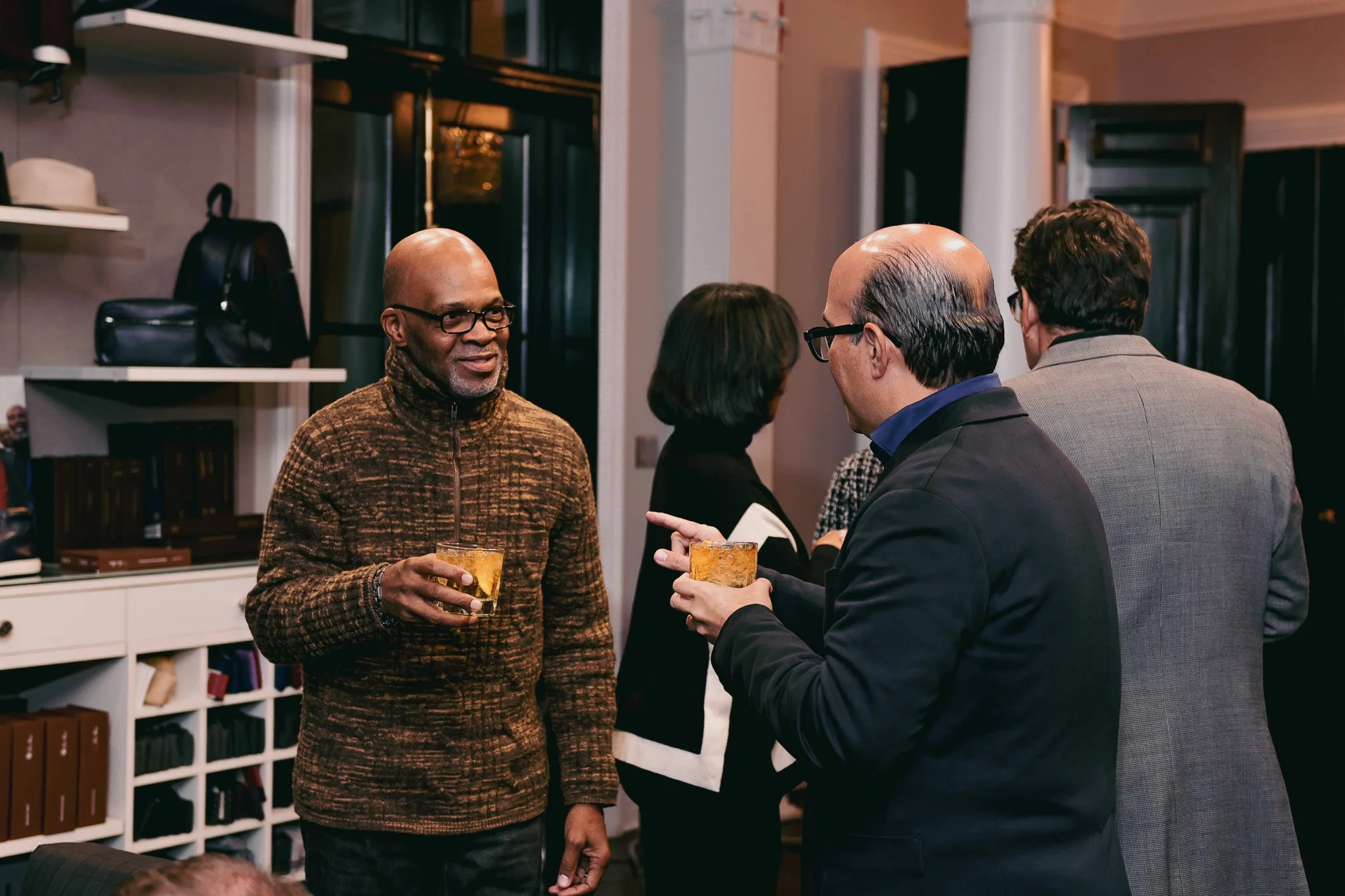 Four adults at a social gathering in a room, two men and two women, holding glasses of drinks and engaging in conversation.