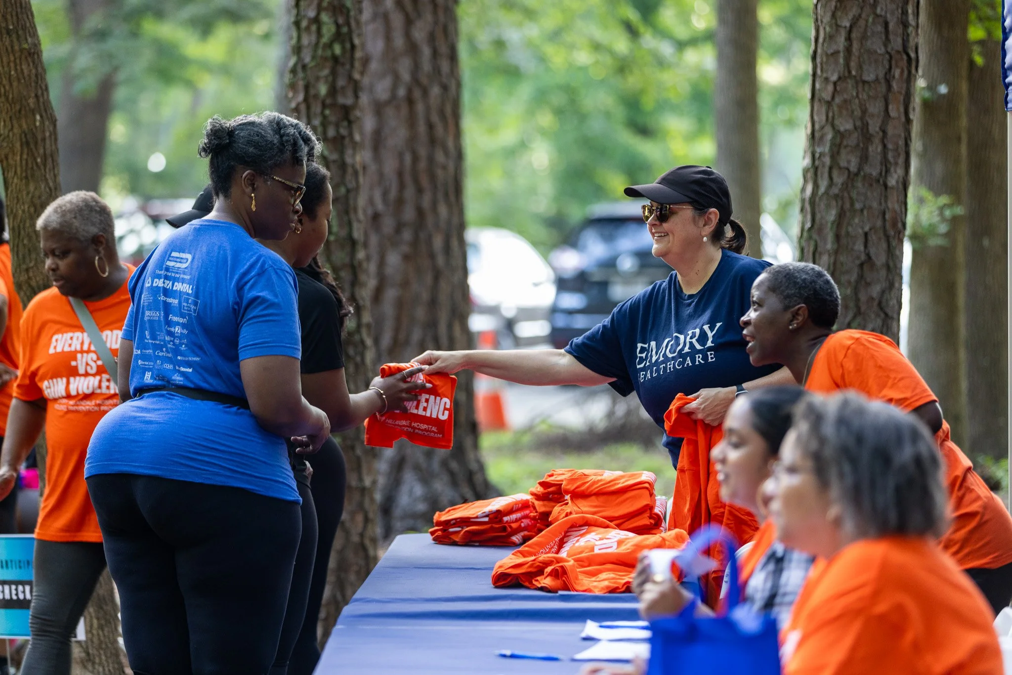 People participating in an outdoor event under trees, with tables displaying orange T-shirts and handing out items, some wearing event shirts and medals.