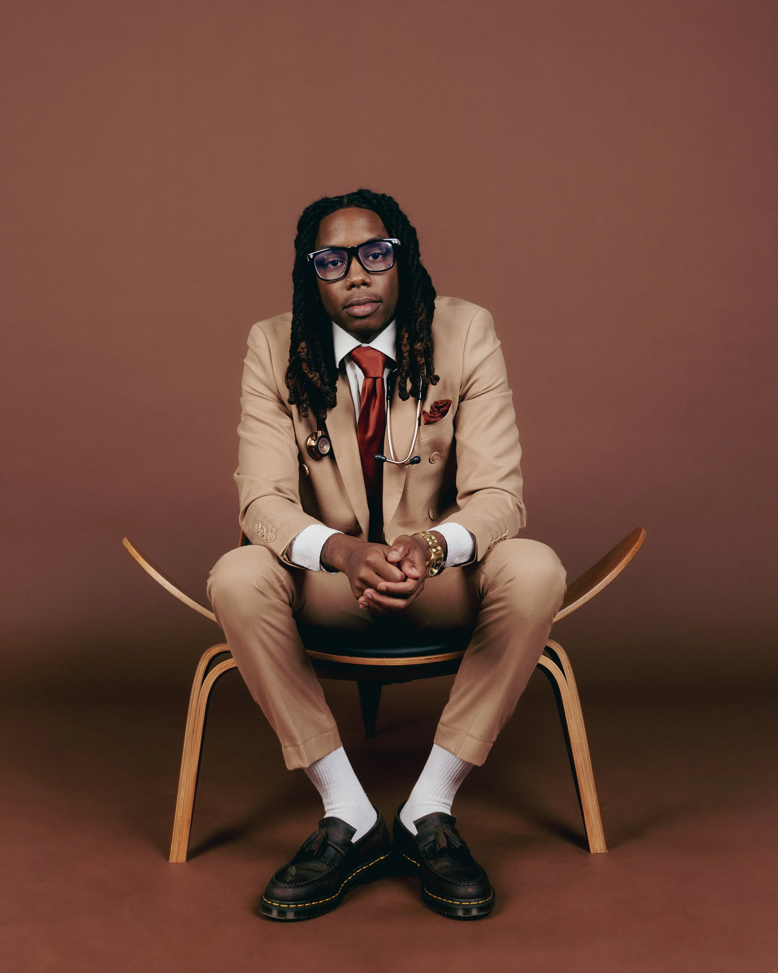 A man seated on a wooden chair in front of a brown background. He is wearing a tan suit, white shirt, brown tie, glasses, and has a stethoscope around his neck. He has long dreadlocks, a gold watch, and black shoes with white socks.