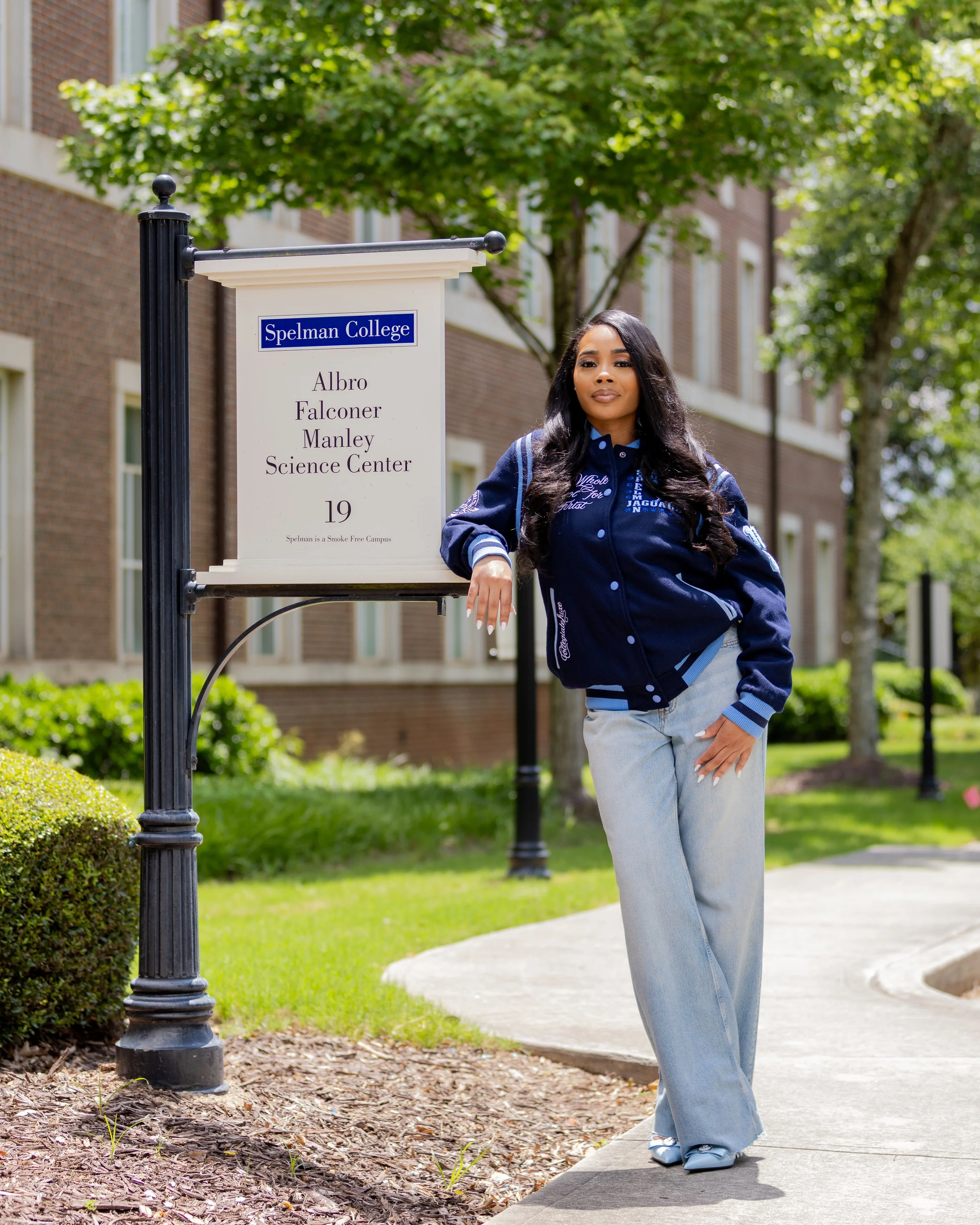 A young woman with long black hair and a confident expression leaning on a sign that reads 'Spelman College, Albro Falconer Manley Science Center, 19, Spelman is a Smoke Free Campus' on a sunny day outside a brick building with green trees and bushes
