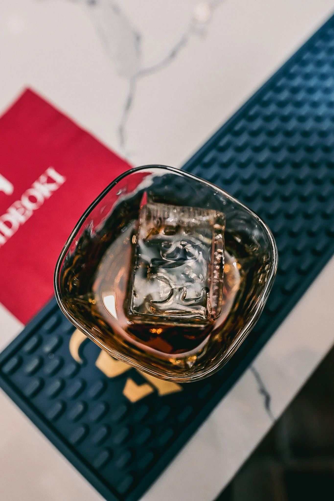 A glass of dark carbonated beverage with ice cubes on a black rubber bar mat with gold writing.