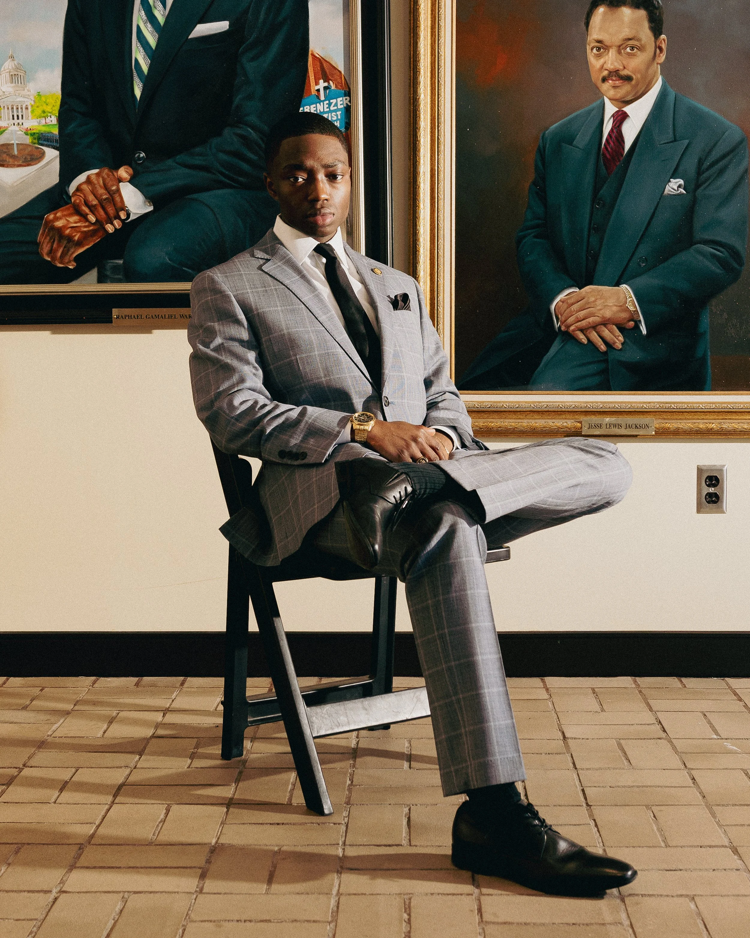 A young man with a serious expression, wearing a light gray plaid suit, white shirt, and black tie, seated on a black chair with his legs crossed, in front of two large portraits in frames. One portrait is of a man with a mustache wearing a dark teal