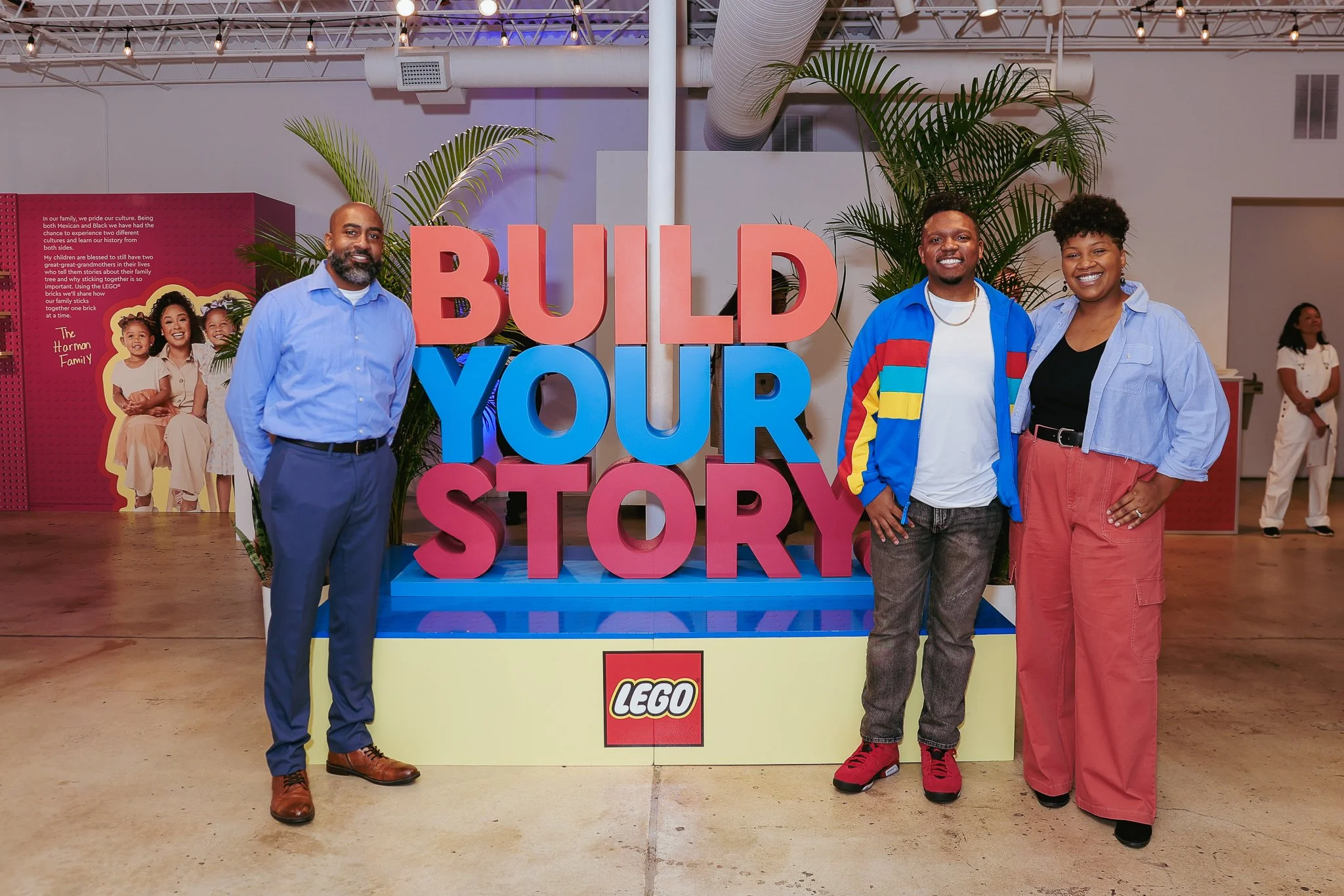Three people standing in front of a large sign that reads 'BUILD YOUR STORY' with a LEGO logo at the bottom. Behind them, there are large green plants and a poster featuring a multigenerational family. The setting appears to be an indoor event or exh