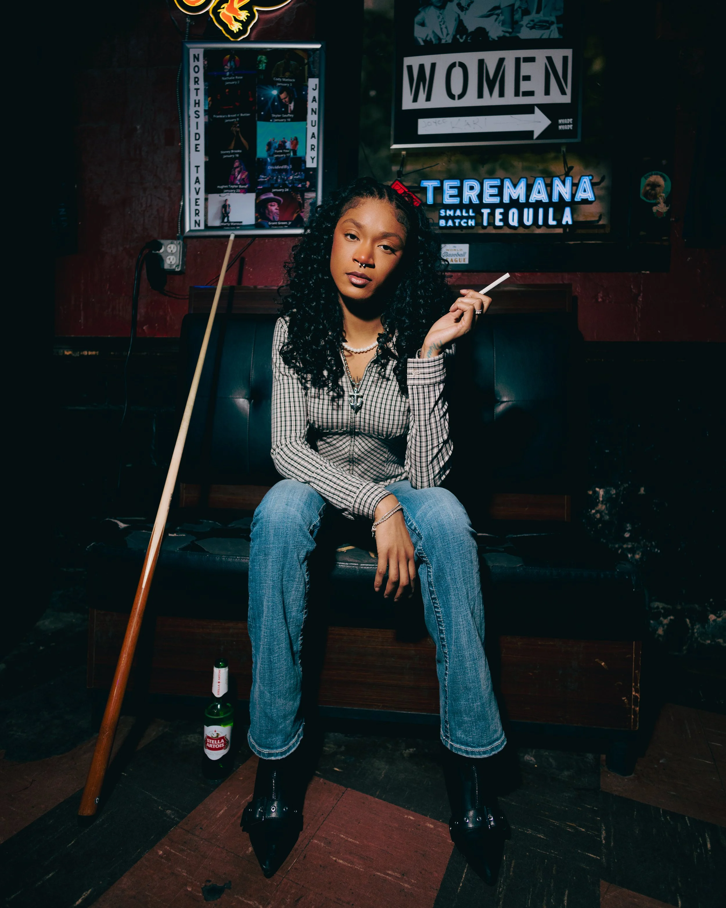 A woman with curly hair and a septum piercing sitting in a dimly lit bar or club, holding a cigarette, with neon signs and posters in the background.
