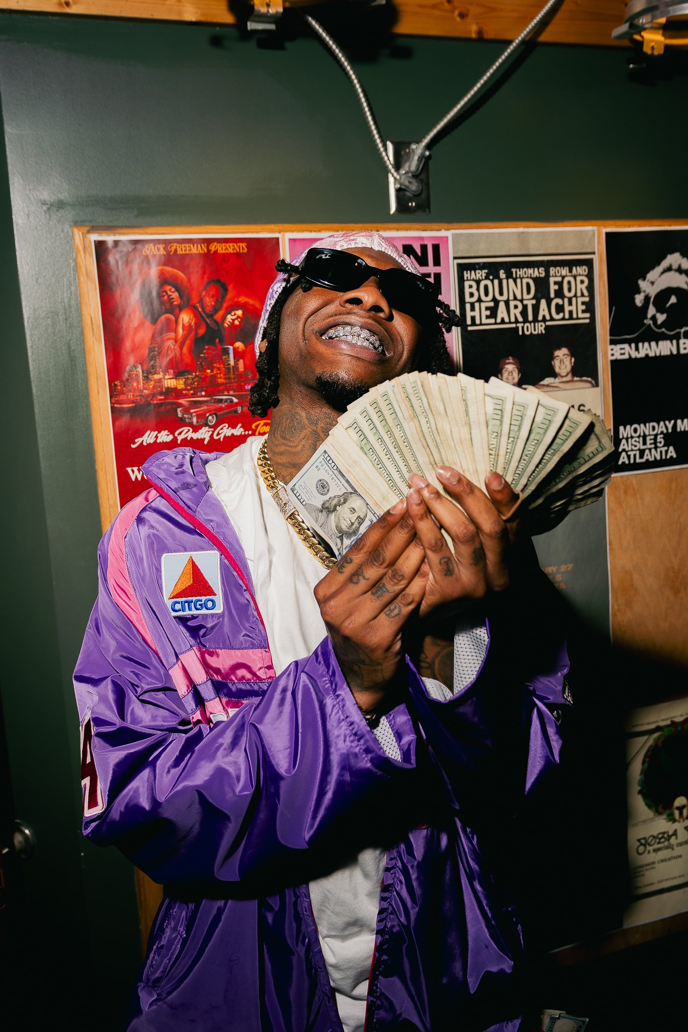A man wearing sunglasses, a purple and pink jacket with a Citgo logo, and a white shirt, holding up a fan of hundred-dollar bills, smiling with grills on his teeth in an indoor setting with posters on the wall behind him.