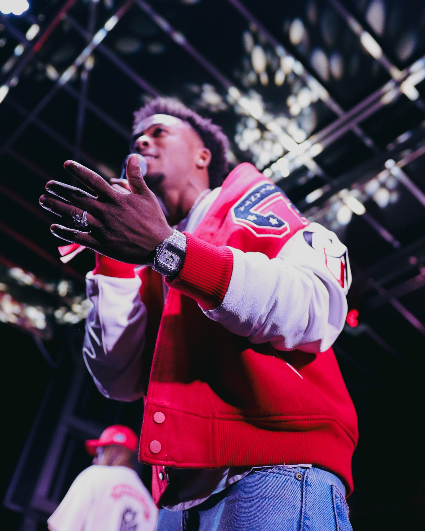 A man with styled hair wearing a red and white varsity jacket, blue jeans, a large watch, and jewelry, speaking or performing with his hand on his chest, against a dark background with bright lights.