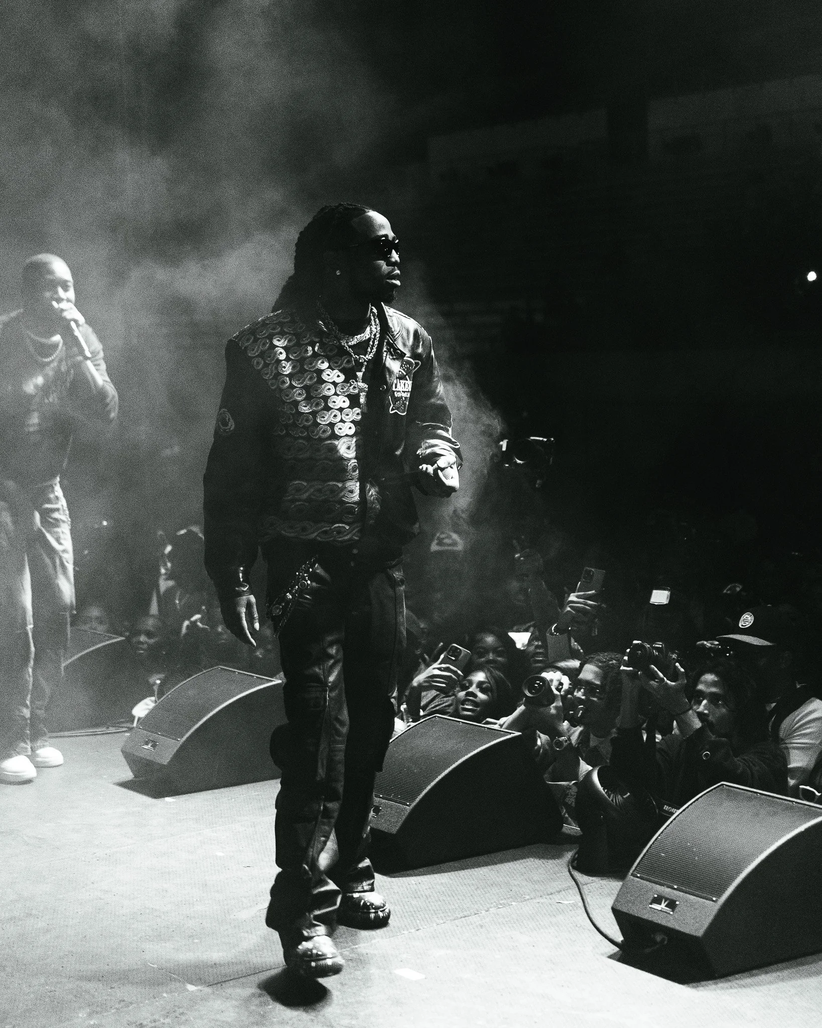 A man wearing sunglasses, a patterned jacket, and multiple chains performs on stage. The crowd is taking pictures and videos with their phones. A woman is on the left side, speaking into a microphone. The stage has speakers and a smoke effect.