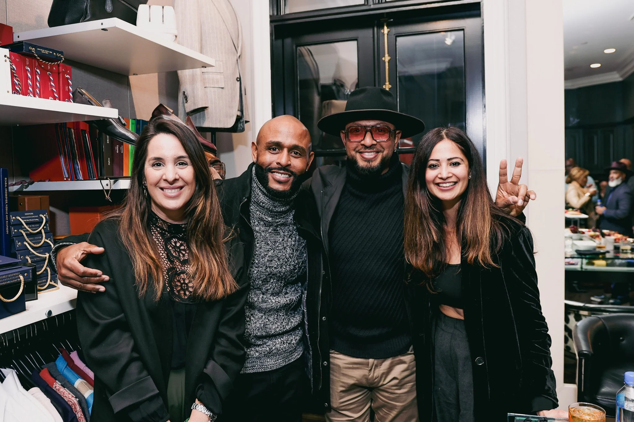 A group of four diverse people smiling and posing for a photo inside a store or boutique. They are standing close together, with a shelf filled with clothing items on their left and a black door behind them.