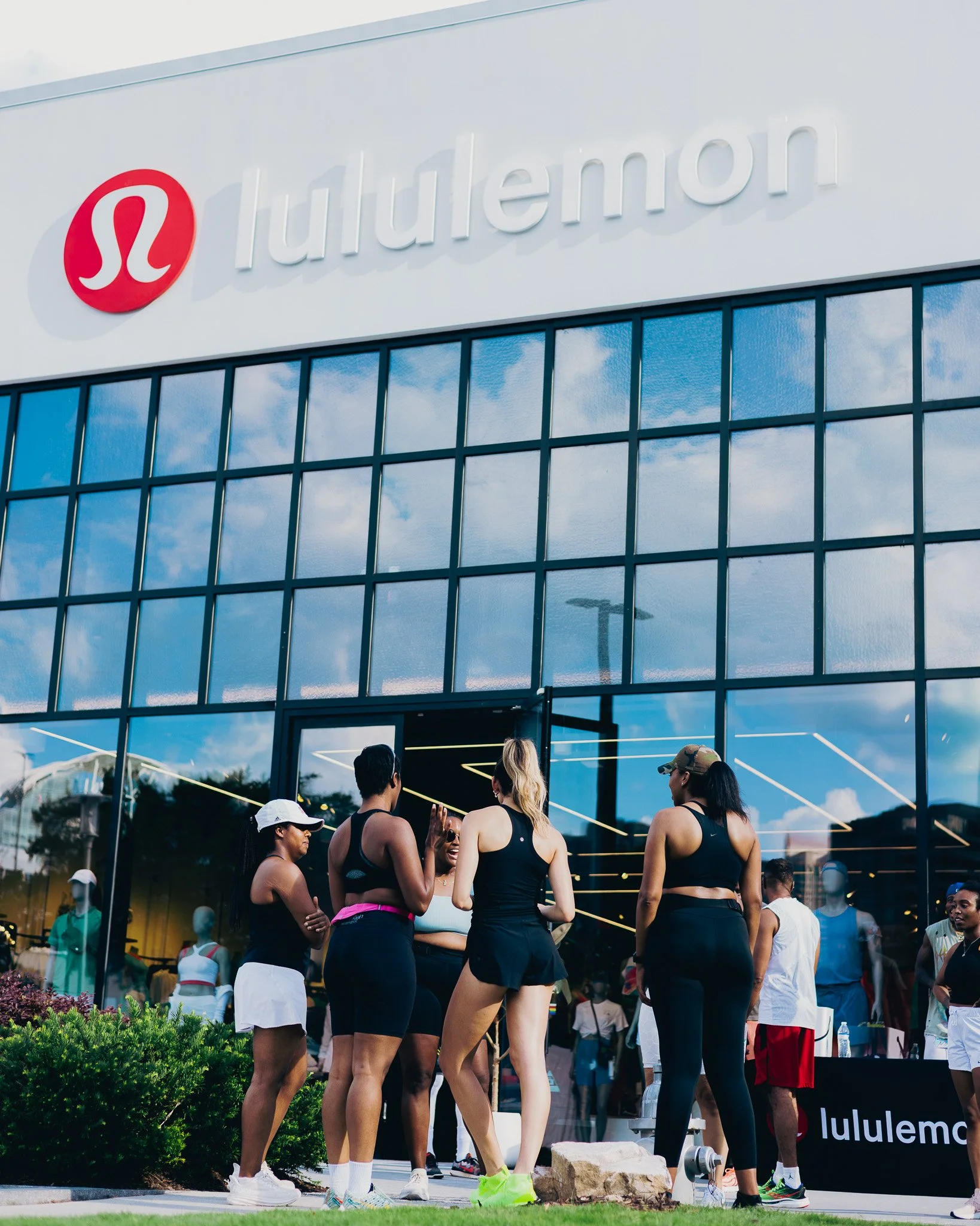 People gathering near a Lululemon storefront with reflective glass windows, preparing for or after a fitness event, with some talking and others wearing athletic clothes.