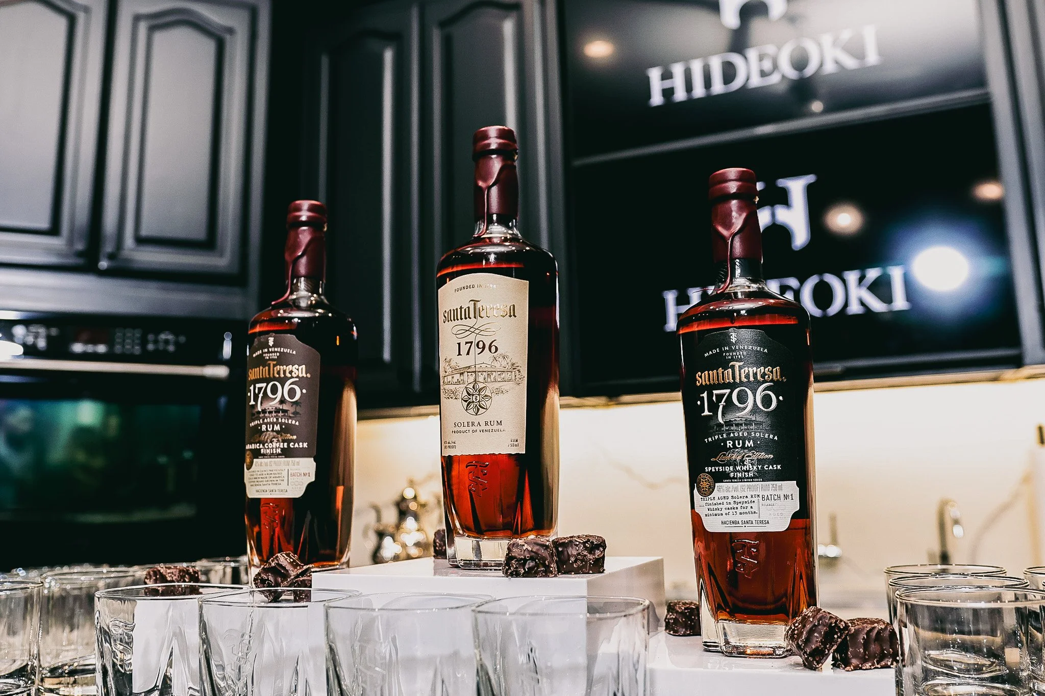 Three bottles of Santa Teresa 1796 rum on display in front of a black cabinet with the brand name 'HIDEOKI' in the background. The bottles are positioned on a white surface with small glasses and chocolate squares around them.