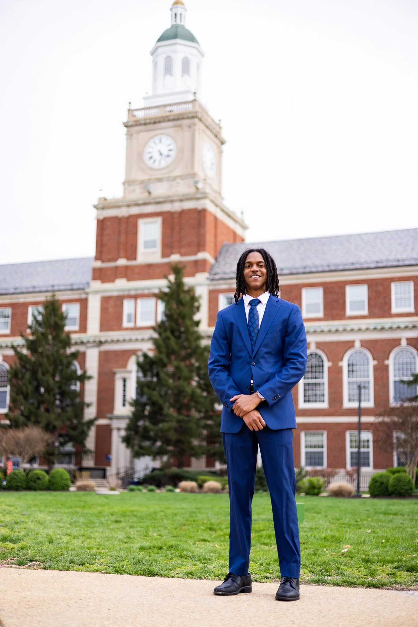 A man in a blue suit standing outside in front of a historic brick building with a clock tower. The man is smiling and has long dreadlocks, with trees and a landscaped area in the background.