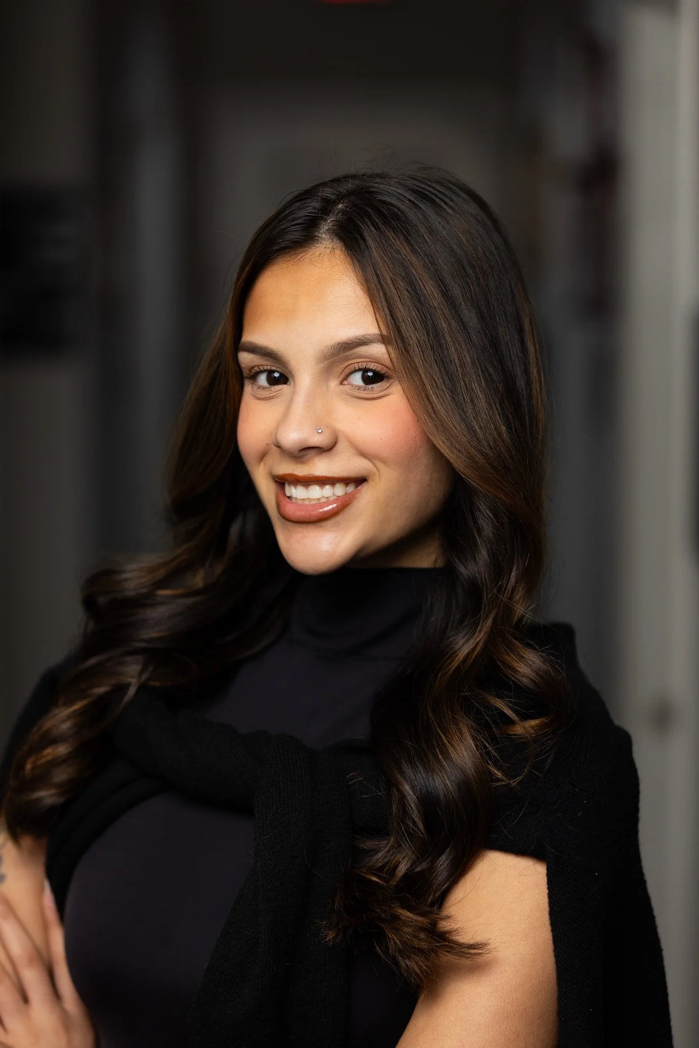 Close-up of a smiling young woman with long, dark wavy hair, wearing a black top, standing against a blurred dark background.