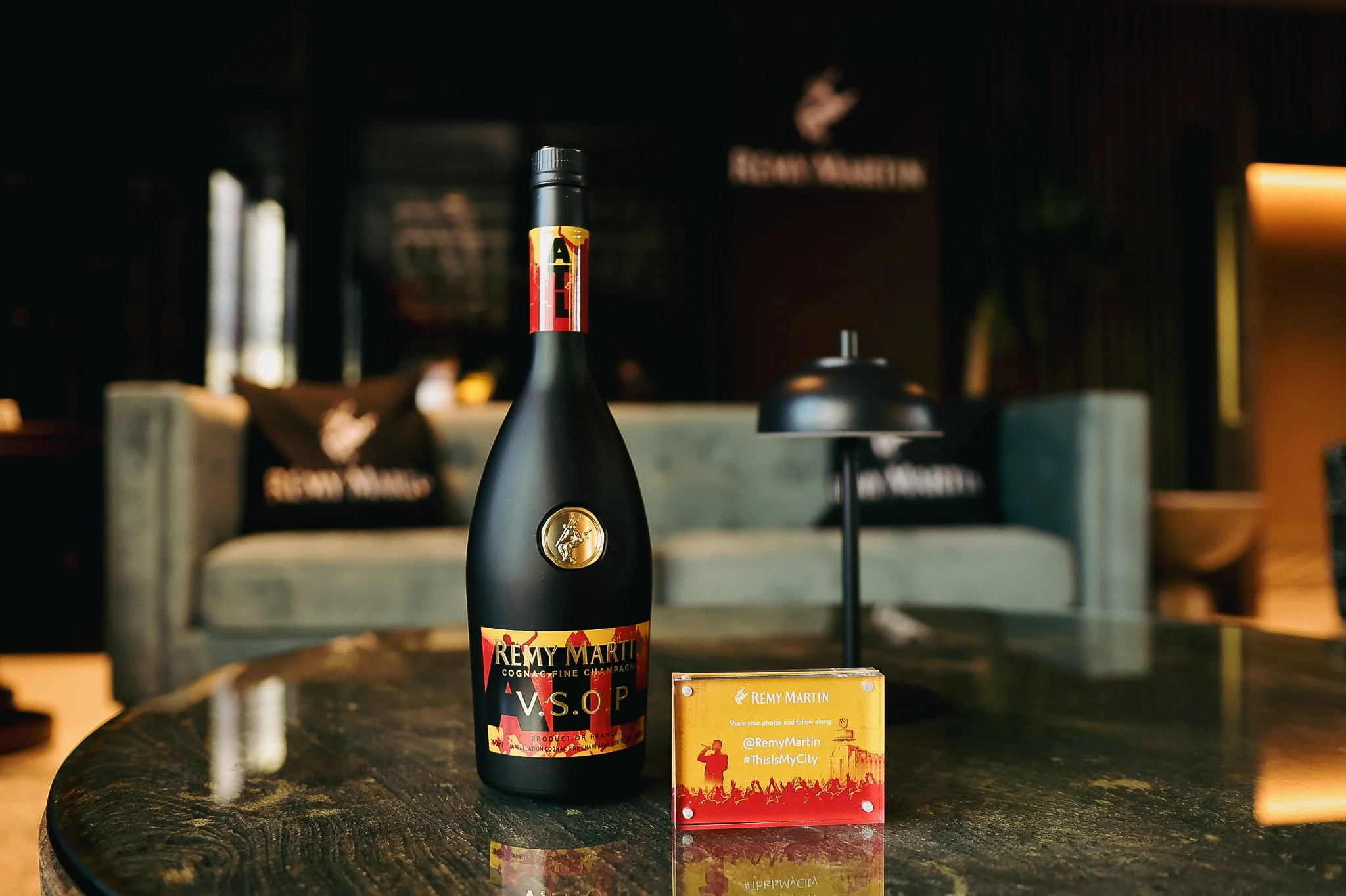Bottle of Rémy Martin V.S.O.P. Cognac on a dark, polished table with a small promotional sign beside it, inside a dimly lit lounge or bar area.